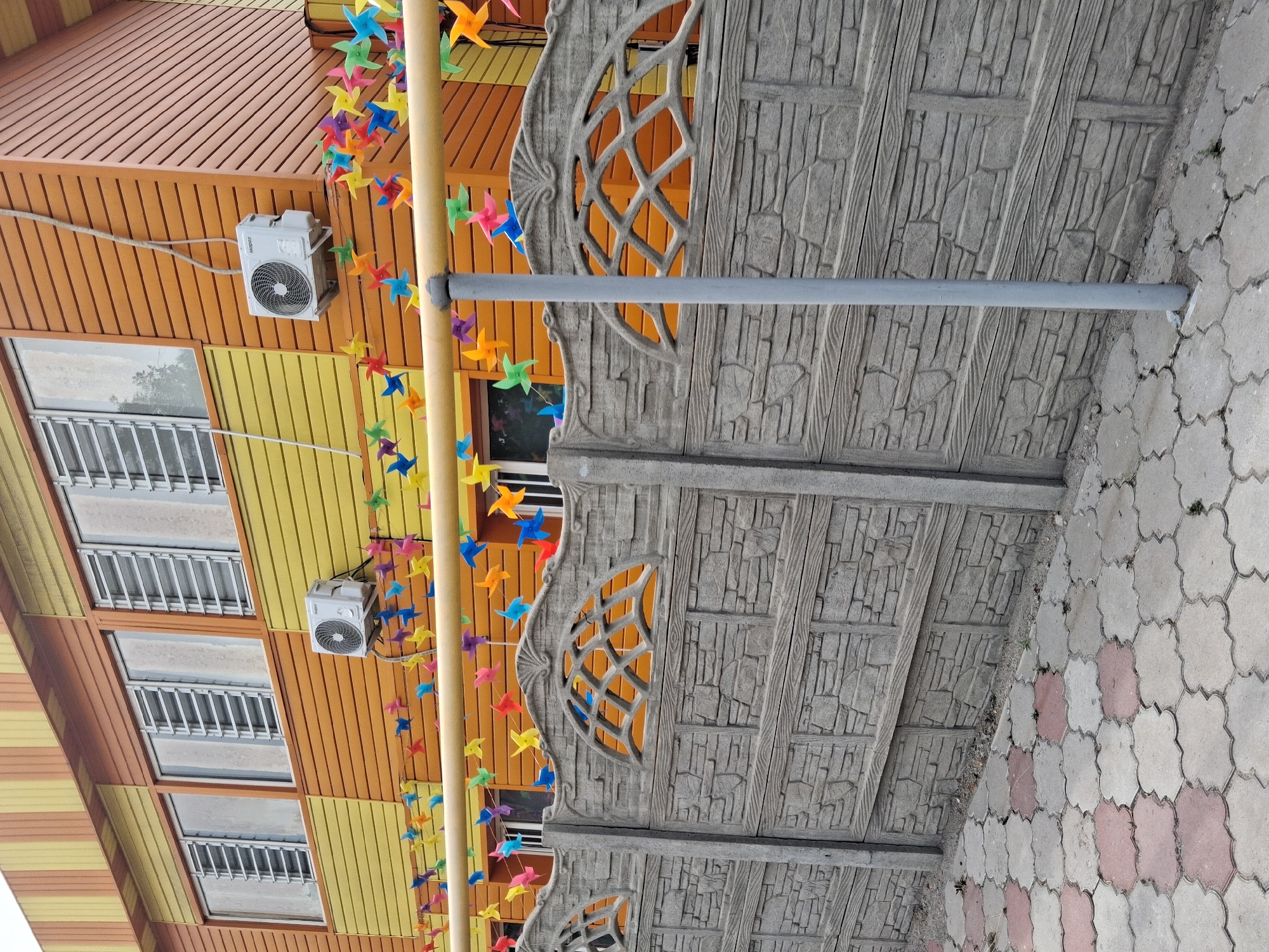 This image depicts the exterior of a building in Guldala, Kazakhstan. The building features a colorful facade with horizontal siding in shades of orange and yellow. Several windows with metal bars are visible on the upper floors. An air conditioning unit is mounted on the orange siding.
A decorative element consisting of a string of colorful pinwheels drapes across the front of the building, adding a playful and festive touch. Adjacent to the building is a gray concrete fence with a textured, stone-like pattern and decorative arched openings.
The ground is paved with interlocking concrete tiles in a light gray and pinkish-red pattern. A thin gray pipe runs horizontally across the frame, possibly part of the building's infrastructure. The scene appears to be outdoors during the daytime, with no specific indications of weather conditions. The overall impression is a bright and somewhat whimsical architectural setting. No people are visible in the image. There is no visible text.