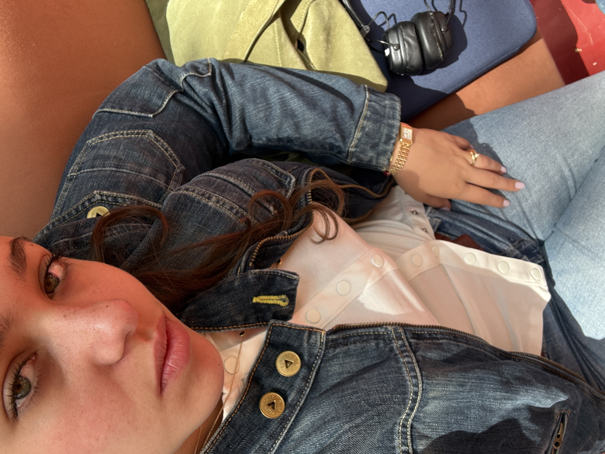 A woman lies on a reddish-brown surface, possibly a couch or a large cushion, with her head close to the camera and her body angled away. She has fair skin, brown hair, and green eyes. She is wearing a denim jacket over a white top with snap closures. A gold watch is visible on her left wrist, and she wears a ring on her right hand. Adjacent to the woman, on the reddish-brown surface, are a lime green bag, and a navy blue object with black headphones on top. The image appears to be a selfie taken from a close, overhead perspective. There are no clear indicators in the image that explicitly confirm the location as Yerevan, Armenia.