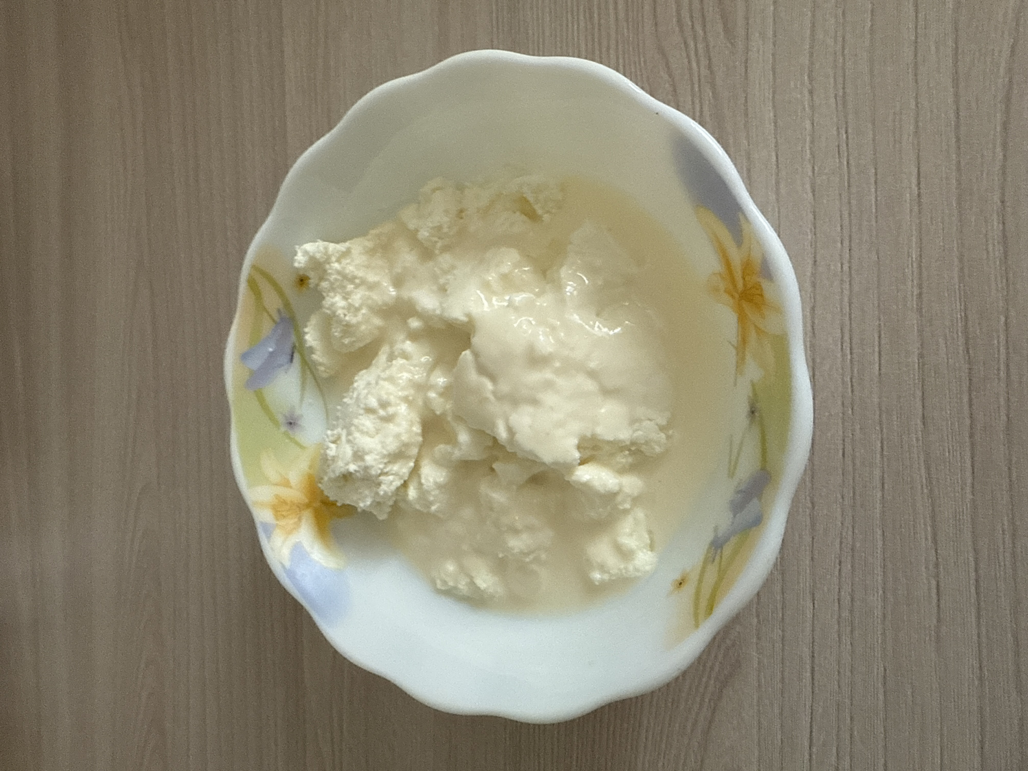 A white bowl with a floral pattern contains a dollop of cottage cheese. The bowl is placed on a wooden surface.