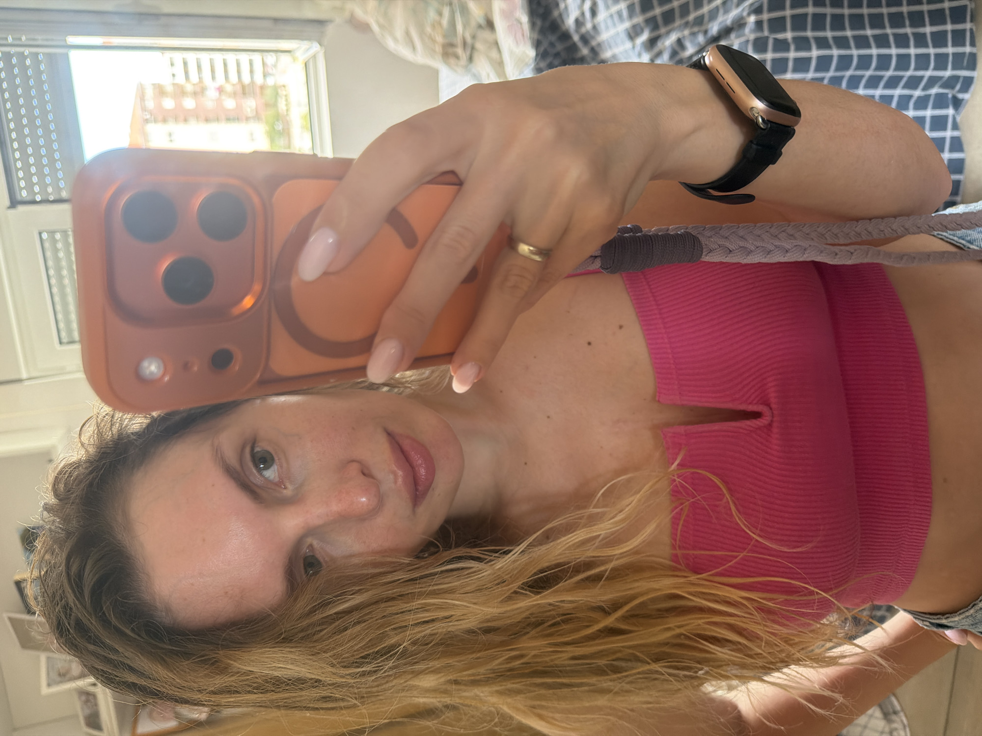 A young woman with long, wavy blonde hair takes a mirror selfie indoors, likely in Budva, Montenegro. She has light skin with freckles, light-colored eyes, and her lips appear slightly glossy. She wears a ribbed, bright pink crop top with a small V-neck cutout and what appears to be denim bottoms.
On her left ring finger, she wears a simple gold band. Her nails are oval-shaped with a light pink or nude polish. On her right wrist, she has a rose gold smartwatch with a black band. She holds an orange-cased smartphone with a prominent triple-lens camera module, and a braided grey strap or lanyard is attached to the phone case.
The background shows an indoor setting, possibly a bedroom or living area, with a blue and white checkered pattern visible, likely from a bed or couch. To her left, a bright window with blinds or shutters reveals an outdoor view of distant buildings, some with reddish or orange roofs, suggesting a residential area. The scene is illuminated by bright daylight, indicating a sunny day. No visible text is present.