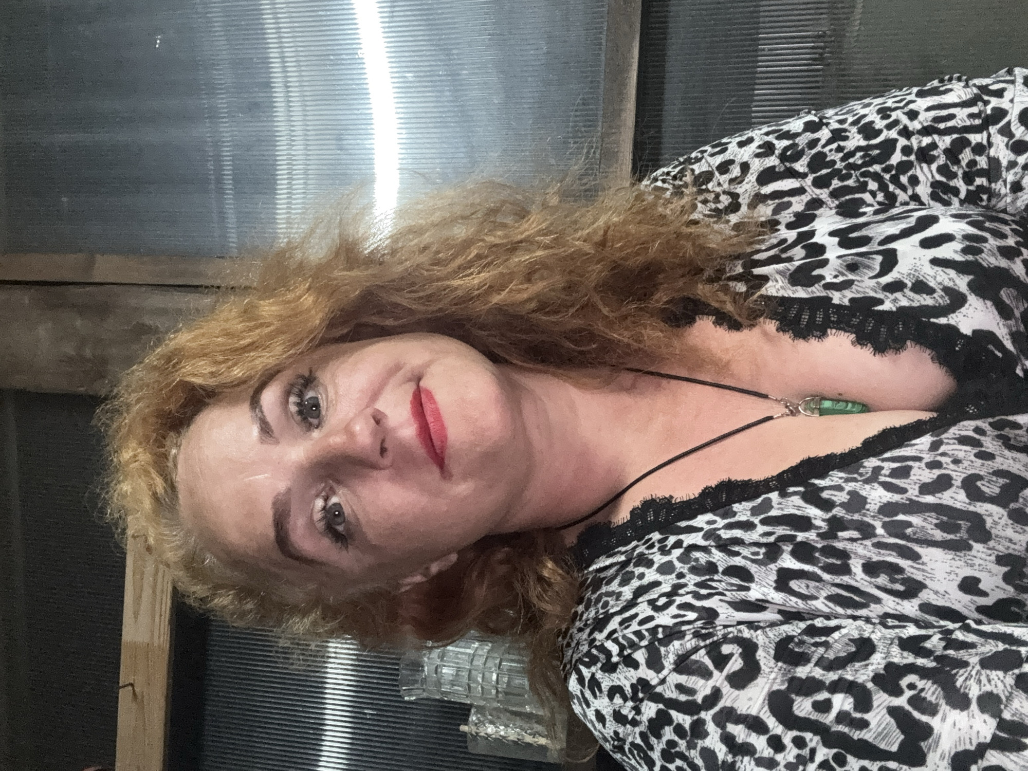 A woman with shoulder-length curly blonde hair and heavy eye makeup is looking towards the camera. She is wearing a black and white leopard print top with black lace trim. A green pendant hangs from a black cord around her neck. She is wearing red lipstick and has a slight smile. The background appears to be a corrugated metal wall with a wooden beam across the top. There are some glass objects on a shelf to the left of the woman. The scene is dimly lit, with light reflecting off the corrugated wall. The location is not identifiable as Chaikovsky, Russia.