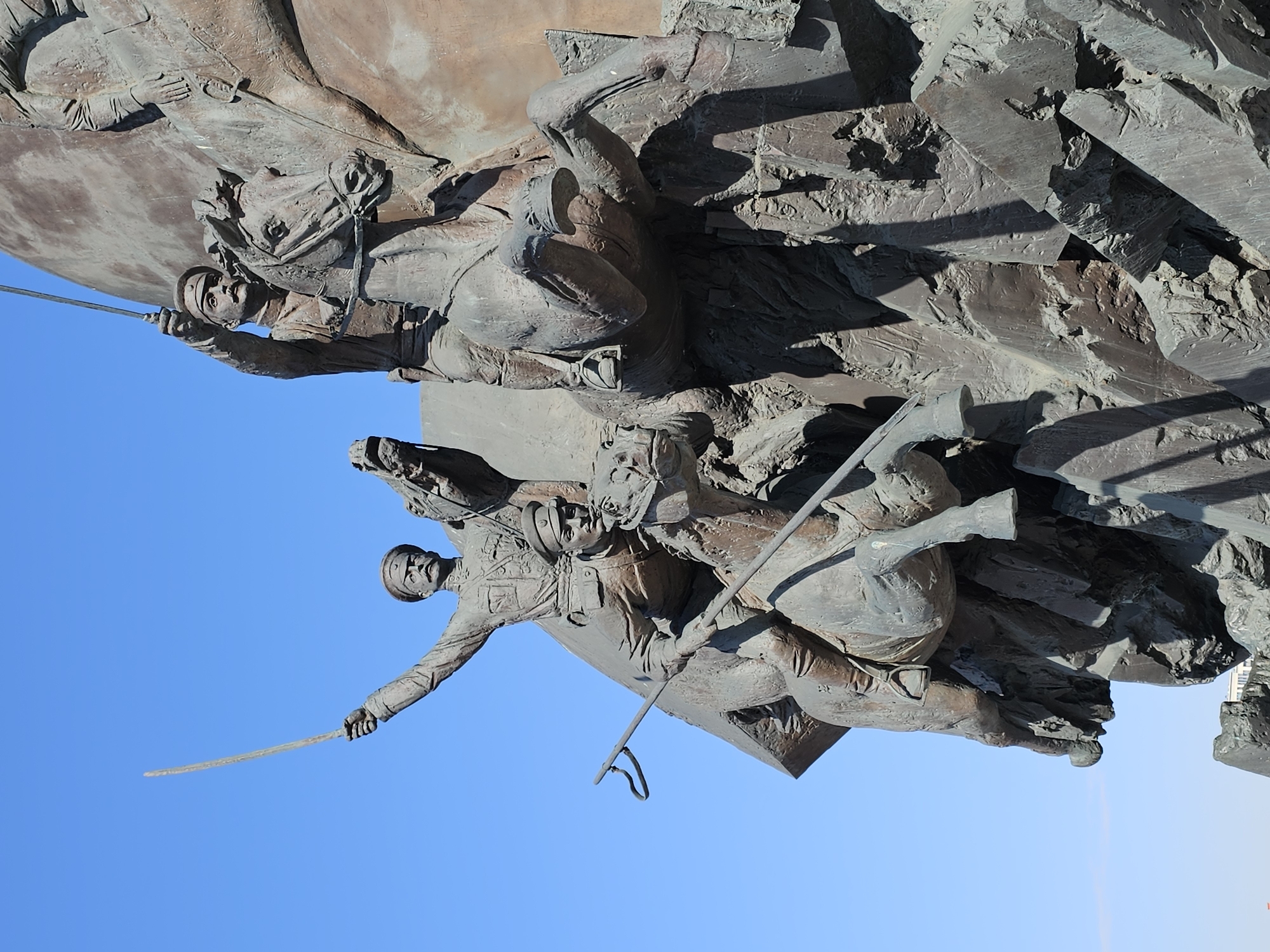 The image shows a bronze relief sculpture of two mounted soldiers. The lower soldier holds a sword and is in motion, while the upper soldier holds a flag. The background behind the soldiers is the blue sky. The location is likely Moscow, Russia, where similar sculptures are found.