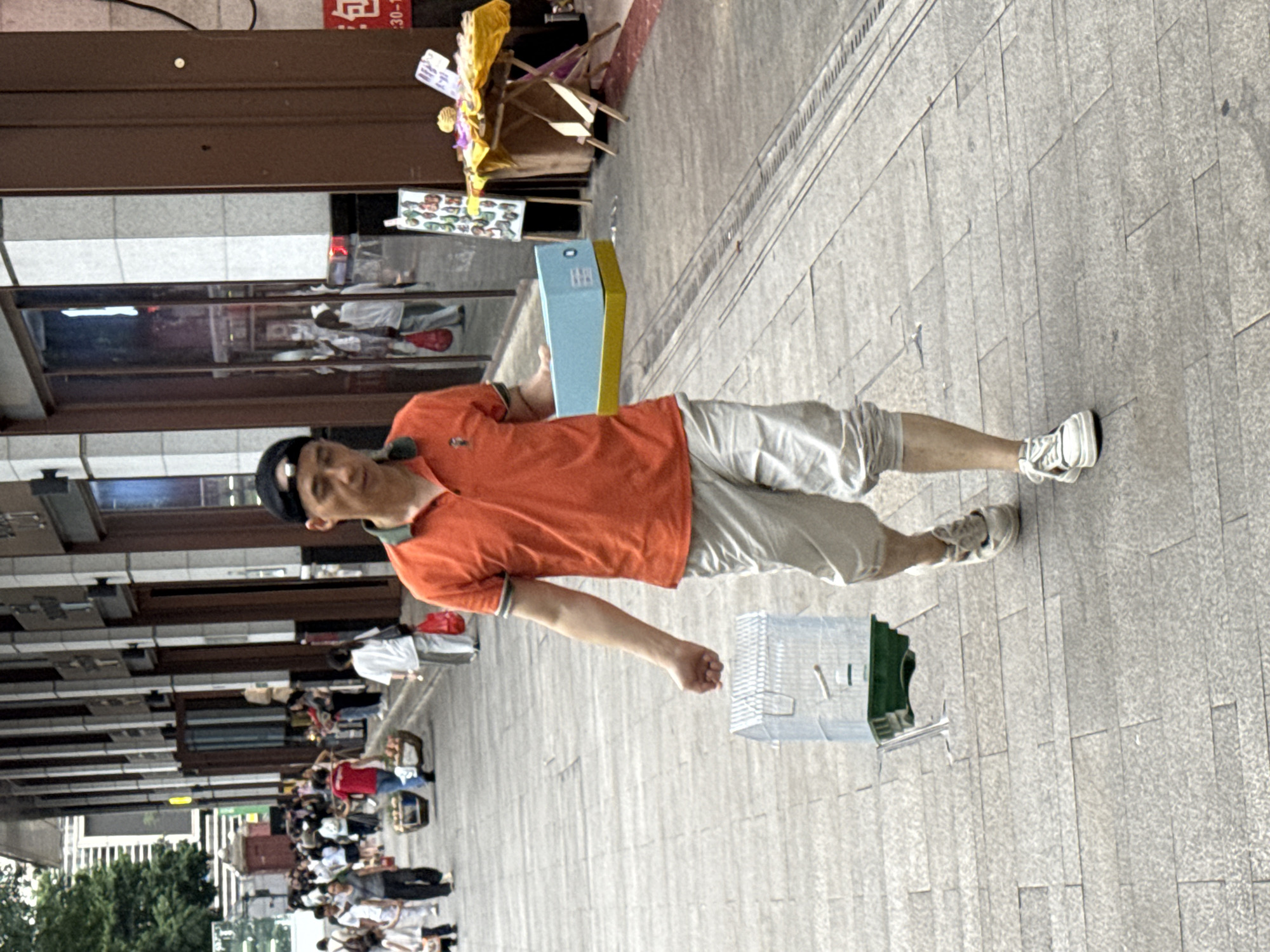A man in an orange polo shirt and light-colored shorts walks along a paved sidewalk in Yuzhong District, China. He is carrying a light blue and yellow box in his right hand and a birdcage in his left. The birdcage is clear plastic with a green base. Behind him, other people are walking in both directions. To his right, a small table with yellow and purple items is set up. The buildings lining the sidewalk have large glass windows and dark brown trim. The sky appears overcast.