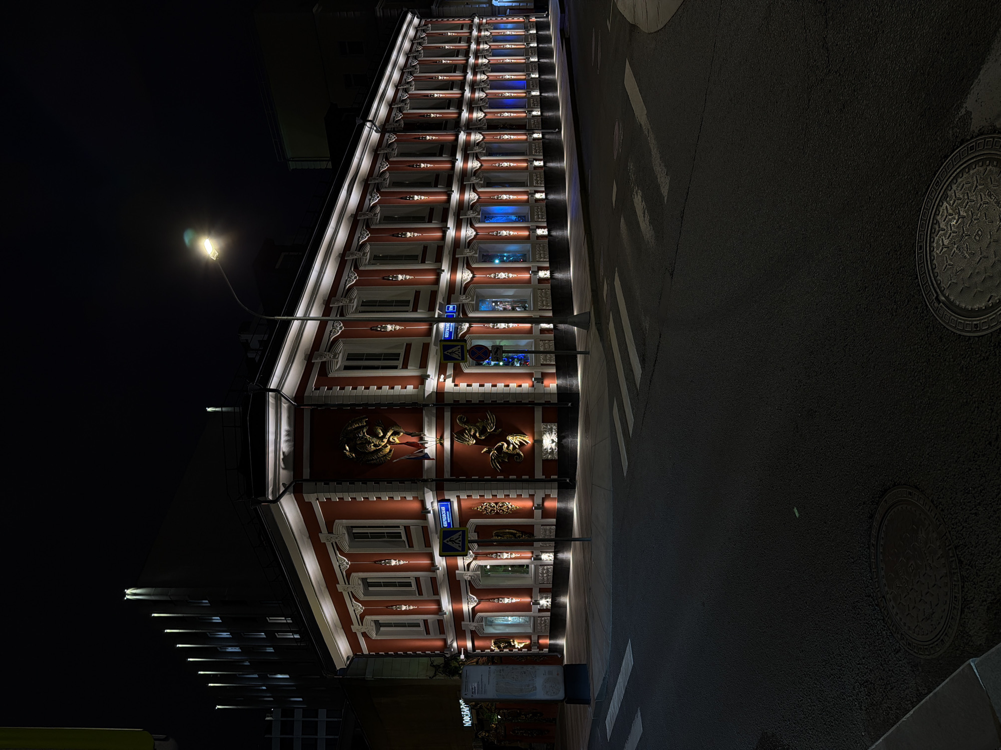 The image depicts a nighttime view of the V. Morozov House in Moscow, Russia, specifically at Podsosensky Pereulok 21. The building, rendered in a predominantly red color palette with white accents, is illuminated by exterior lighting, emphasizing its architectural details, including ornate window frames and golden dragon sculptures. The building's facade is visible along the left side, with additional buildings and streetlight noticeable in the background. A street runs along the right side of the image, and features a pedestrian crossing and a manhole cover. The lighting suggests the time of the photograph is dusk or night. There are no visible people.