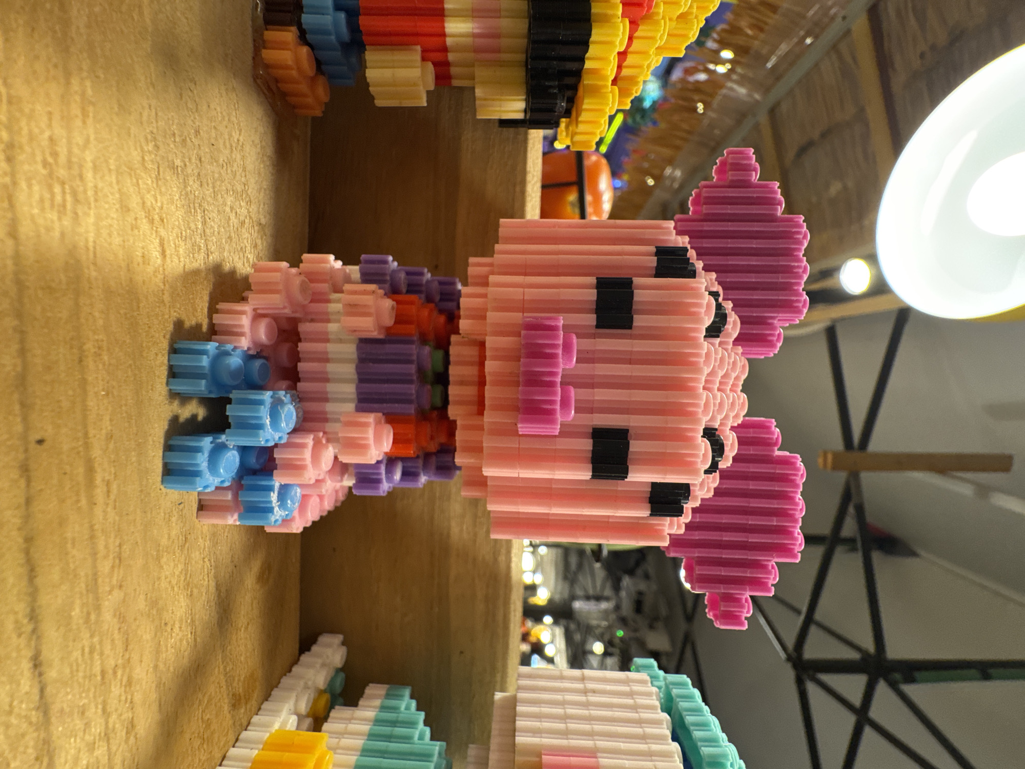 This image, taken in Karon, Thailand, shows a close-up view of a pink pig figurine constructed from interlocking plastic bricks. The figurine is sitting on a wooden surface, with other brick constructions visible on either side, including a yellow and red stack to the left and a turquoise and white stack to the right. The pig figurine has large pink ears, black eyes, and a pink snout. It is wearing a colorful outfit made of purple, pink, and white bricks. The background is blurred, showing a dimly lit indoor environment with what appears to be part of a ceiling fixture and structural elements. The lighting in the image suggests an artificial light source, likely from above, casting shadows on the wooden surface.