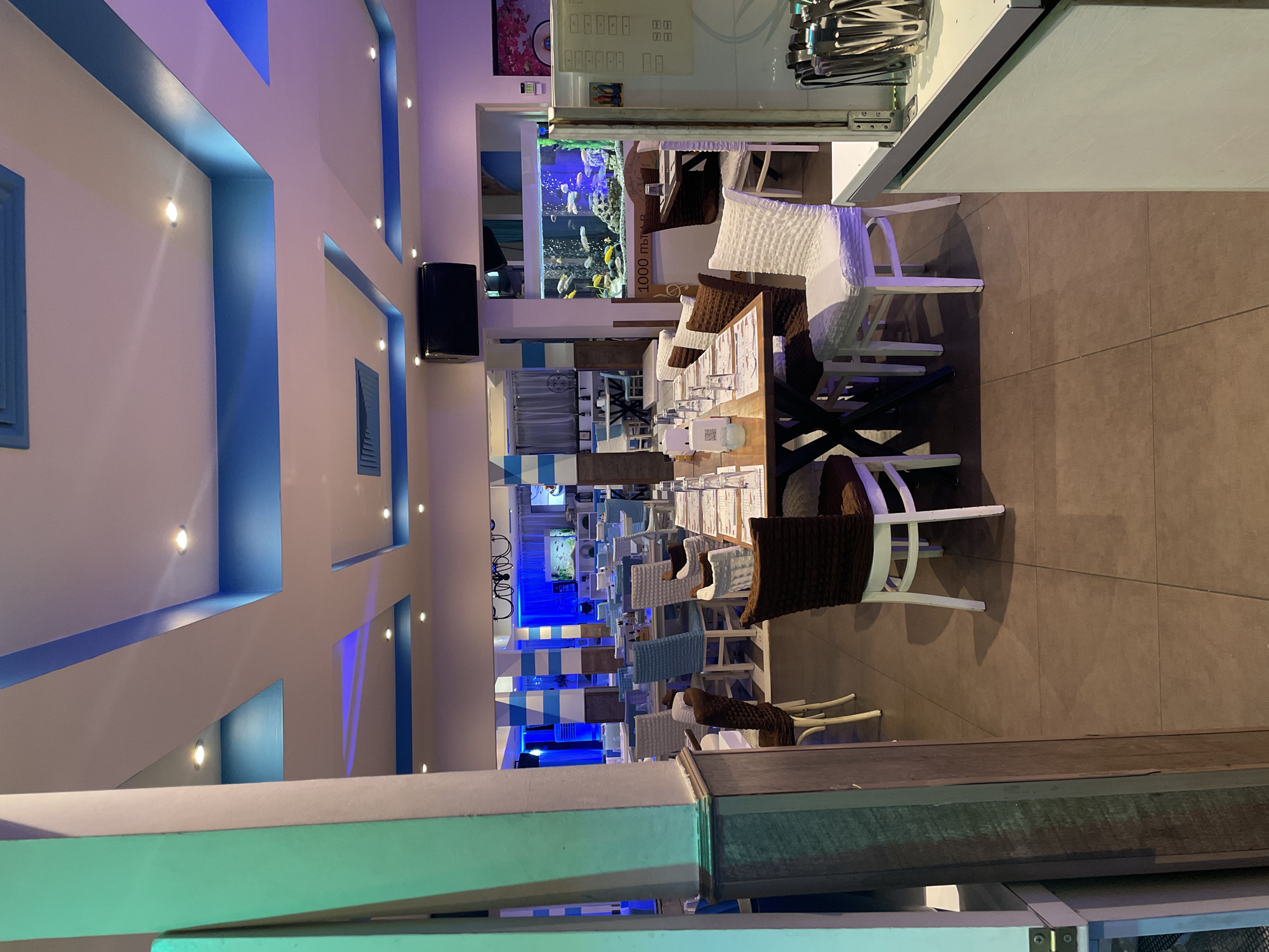 This image depicts the interior of an empty restaurant or cafe, likely set for service, located in Plovdiv, Bulgaria. The establishment showcases a distinctive blue and white color palette, evoking a Mediterranean or coastal ambiance.
The space is illuminated by a combination of artificial lighting. White spotlights are integrated into the walls and ceiling, while blue LED strip lighting highlights architectural recesses and decorative elements along the walls, creating a vibrant atmosphere. The walls are mostly white, featuring blue accents that resemble decorative shutters or windows. A black speaker is mounted high on one wall.
The dining area is furnished with numerous tables, each meticulously set with glasses and what appear to be menus or information stands, including small QR code displays. The chairs are a mix of styles, predominantly white woven wicker-like designs, with some having brown cushioned backs, suggesting a comfortable seating arrangement. The floor is tiled with large, rectangular brown tiles.
A prominent and eye-catching feature of the restaurant is a large, well-lit aquarium filled with a variety of colorful fish, serving as a vibrant centerpiece. Another smaller aquarium is visible further into the room. Above a counter or doorway, there is a grid-like display with small, unidentifiable icons.
Visible text includes "1000 пъти в" printed on a decorative pillar next to the main aquarium. "Пъти" translates to "times" in Bulgarian, indicating part of a larger phrase. The absence of people, coupled with the artificial lighting, suggests the image was taken during the evening or night, either before opening or after closing hours. The overall impression is one of a clean, modern, and inviting dining space.