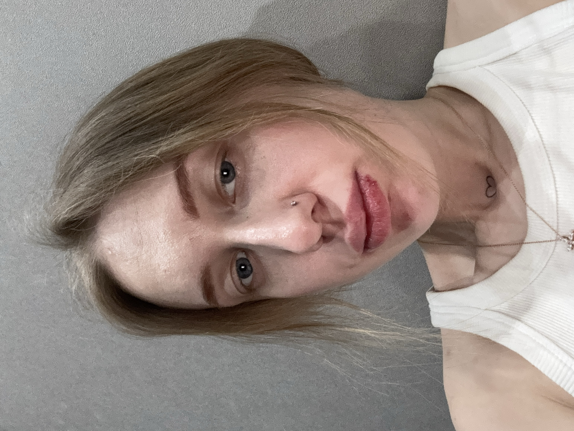 A female subject, appearing to be an adult, is depicted in a close-up, front-facing portrait. She has light-colored hair, blue eyes, and fair skin. A silver stud piercing is present on the right side of her nose. Her lips exhibit injuries: the lower lip shows a dark red lesion, and the upper lip also has reddish discoloration, consistent with trauma or a healing injury. A small, black heart-shaped tattoo is visible on her neck. She is wearing a white ribbed tank top and a gold-colored necklace with a cross pendant. The background consists of a plain, light grey wall with a textured finish. The specific location is Rostov-on-Don, Russia.