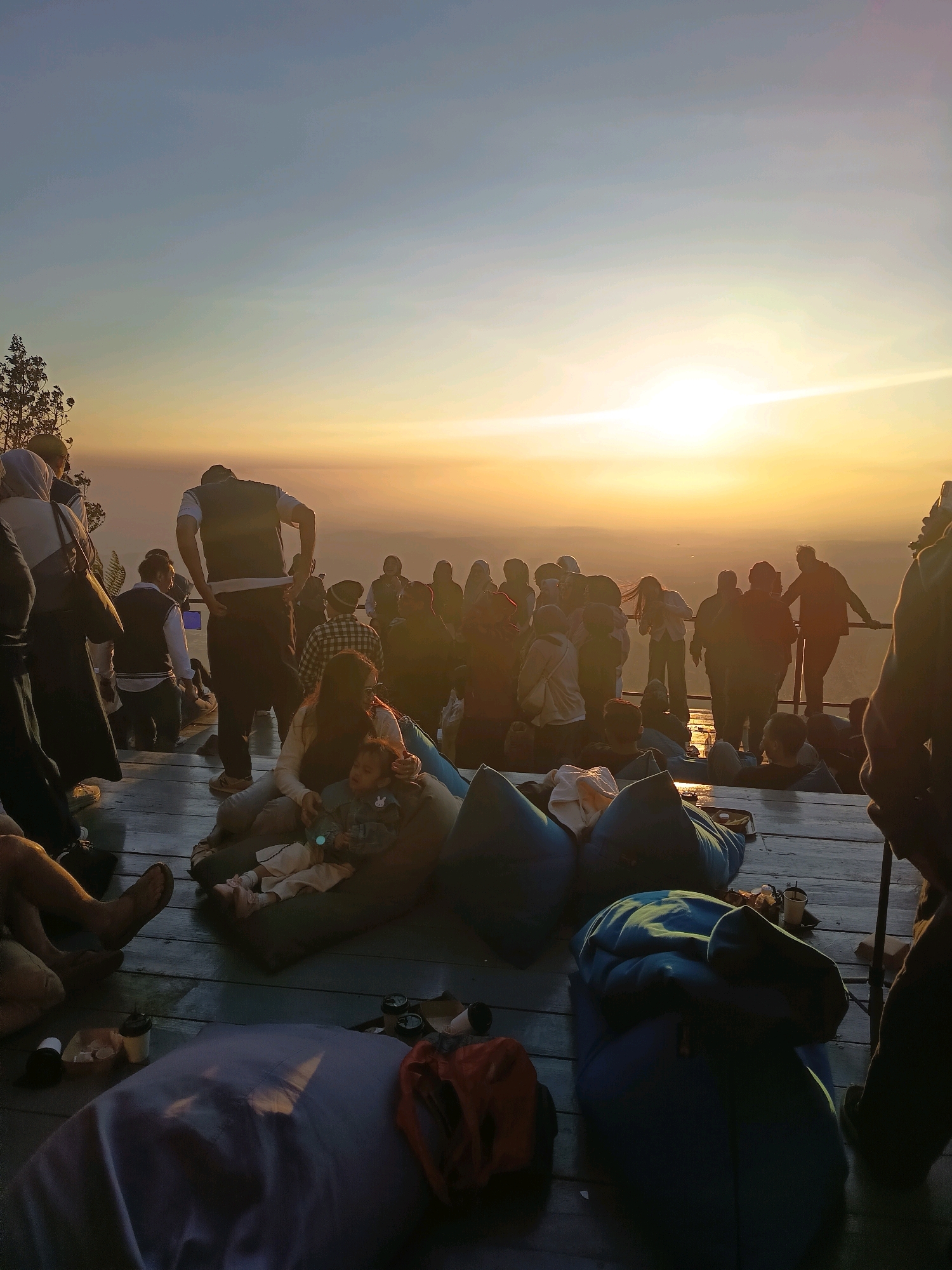 This is a photograph taken from a high vantage point overlooking a vast expanse, likely a mountain or cliffside overlooking a plain or body of water. The time of day appears to be either sunrise or sunset, given the warm, golden light illuminating the sky and creating a hazy glow over the landscape.
In the foreground, a group of people are gathered on a wooden deck, seated on large beanbag chairs and on the deck itself. Some are dressed casually, while others wear headscarves. A woman is holding a small child, who is also seated on a beanbag. There are several cups and other small objects scattered on the deck.
In the mid-ground and background, more people are visible, standing and milling about, also appearing to be enjoying the view. They are silhouetted against the bright sky, making it difficult to discern individual features. The overall atmosphere is one of relaxation and shared enjoyment of the natural scenery. The location is identified as Keditan, Indonesia, suggesting this is a popular scenic spot.