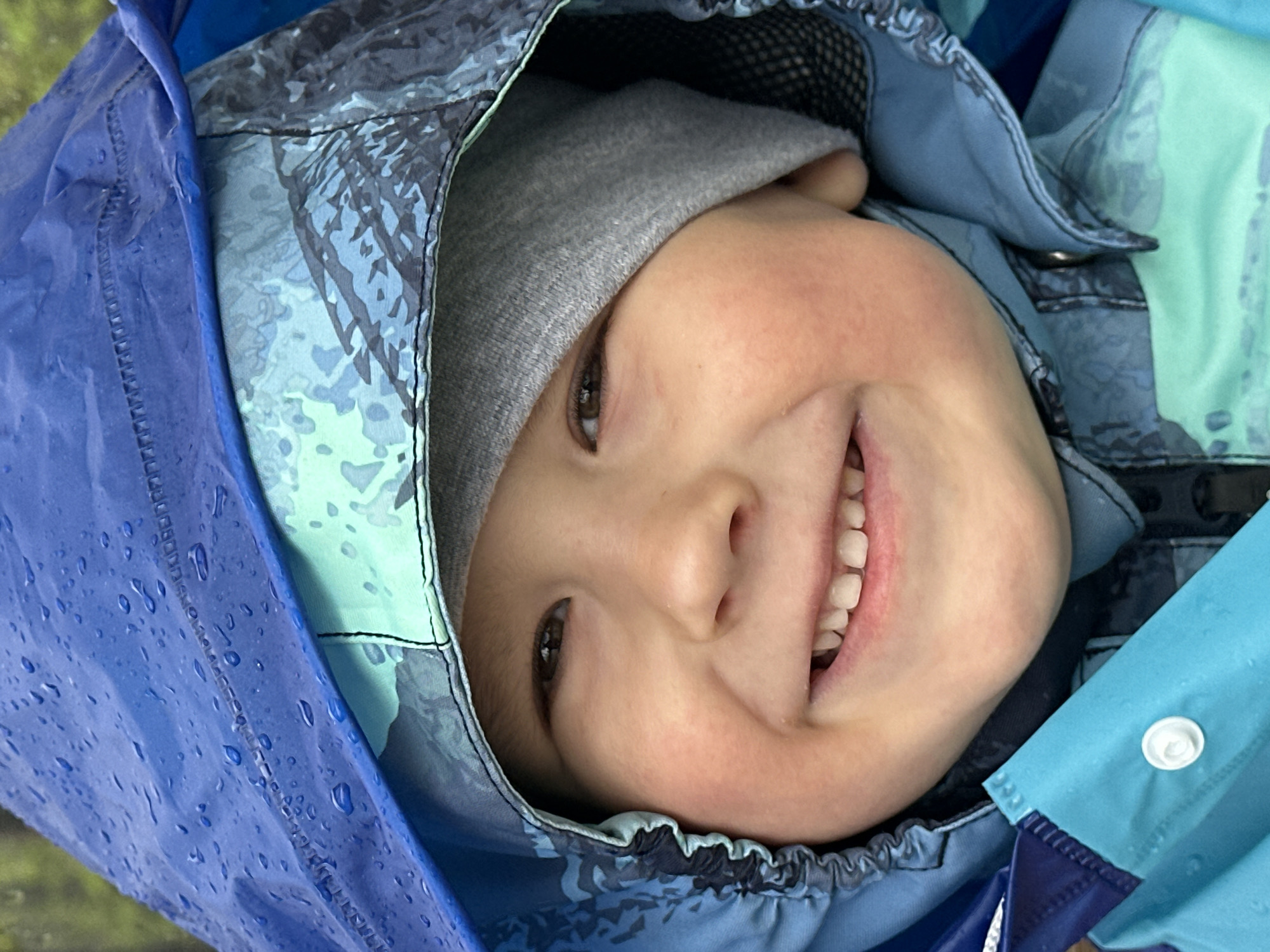 A young child, possibly between 3 and 6 years old, is positioned looking into the camera, seemingly under the hood of a blue and gray camouflage-patterned raincoat. The child also wears a gray beanie cap under the hood. The child is smiling, revealing upper teeth. The hood appears to be slightly wet, with water droplets visible on the blue portion. The photo may have been taken outdoors. There is no clear indication of Nizhny Novgorod, Russia, or any urban environment.
