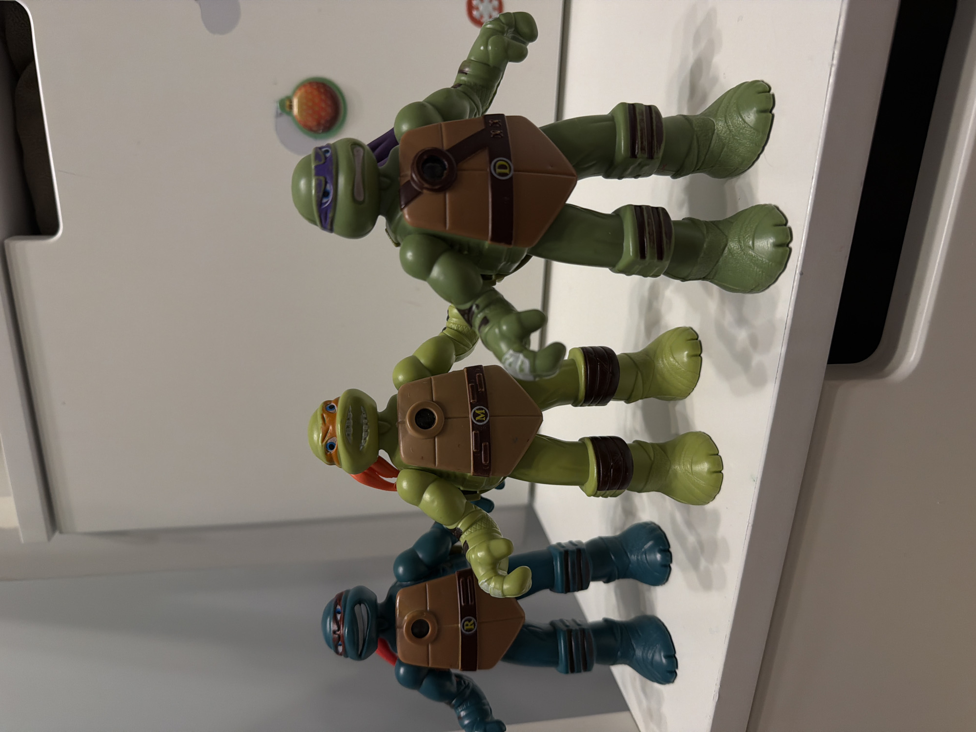 This media file features three Teenage Mutant Ninja Turtles action figures, arranged vertically from top to bottom on a light-colored, flat surface. The image is rotated 90 degrees clockwise, making the figures appear to be lying on their backs with their heads facing to the right.
The top figure, presumably Donatello, is green with a purple mask and a brown shell bearing the letter "D". It has a serious or determined expression. The middle figure, Michelangelo, is a lighter green with an orange mask and an "M" on its brown shell, displaying a wide, open-mouthed smile. The bottom figure, Raphael, is teal/blue with a red mask and an "R" on its brown shell, showing a slight grin. All figures feature textured green or blue skin and wear brown wrist and ankle wraps.
The figures are posed as if standing upright, suggesting they are on display. The setting appears to be indoors, likely on a shelf or cabinet top against a plain white background. There are two small, round, possibly magnetic decorative items visible on the background: an orange and green one near the top figure's head, and a partially visible blue and green one behind the middle figure's head.
No specific activity or event beyond their static display is taking place. The lighting is even, and there are no discernible location cues within the image itself to confirm the provided context of Sofiivska Borshchahivka, Ukraine. The time of day and weather cannot be determined.