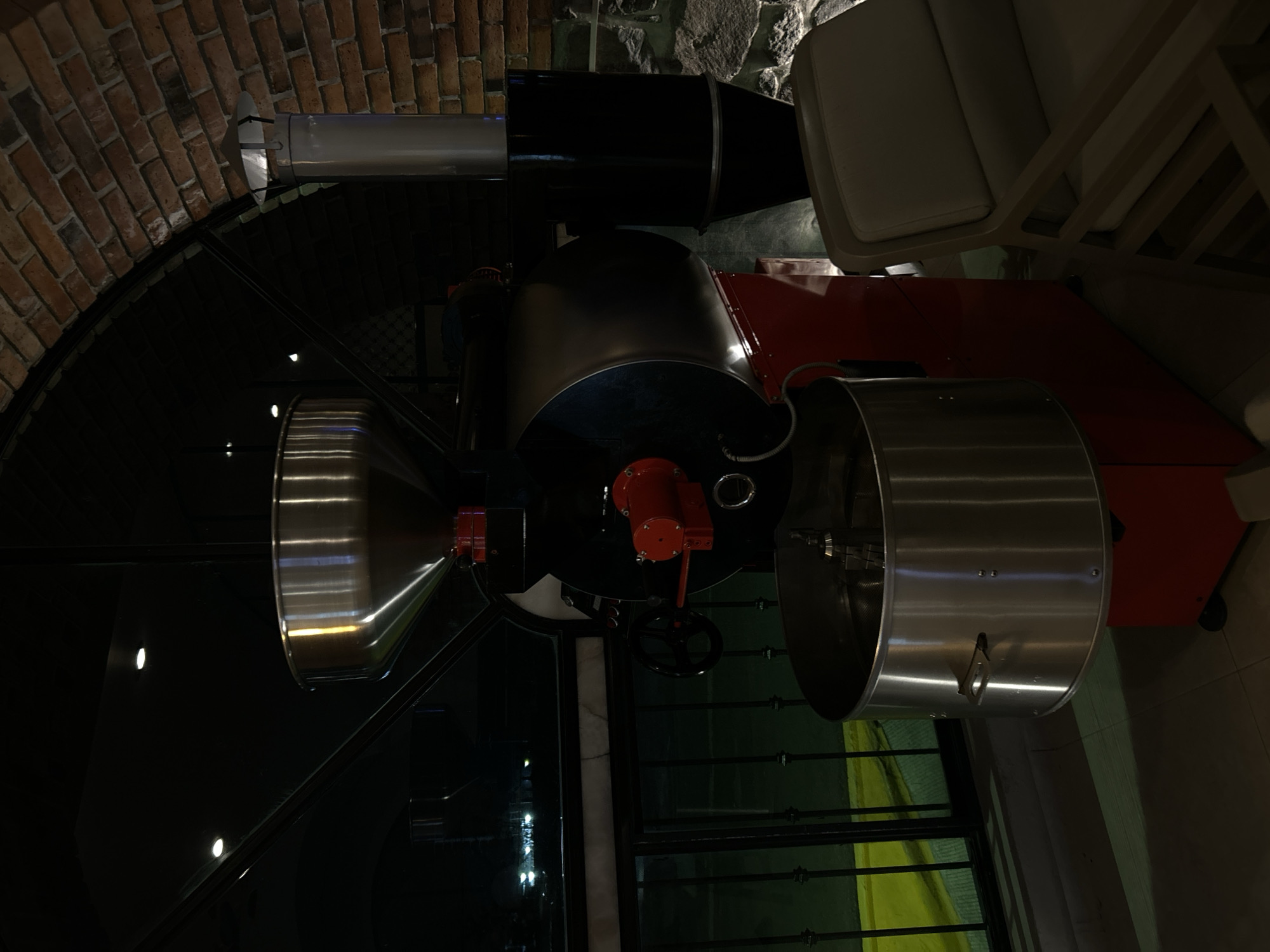 A coffee roasting machine is positioned indoors, presumably in Santa Ana Yancuitlalpan (Chilhuacán), Mexico, as specified. The machine features a large silver funnel on its left, leading into a black cylindrical central section. A red component is visible on this section. To the right, a larger silver cylinder is attached. A black exhaust pipe extends upwards, next to a chair with white cushions. The background shows a brick wall and stone masonry. Below the machine, a window with metal bars is visible, along with a yellow and green curtain. The floor appears to be tiled.