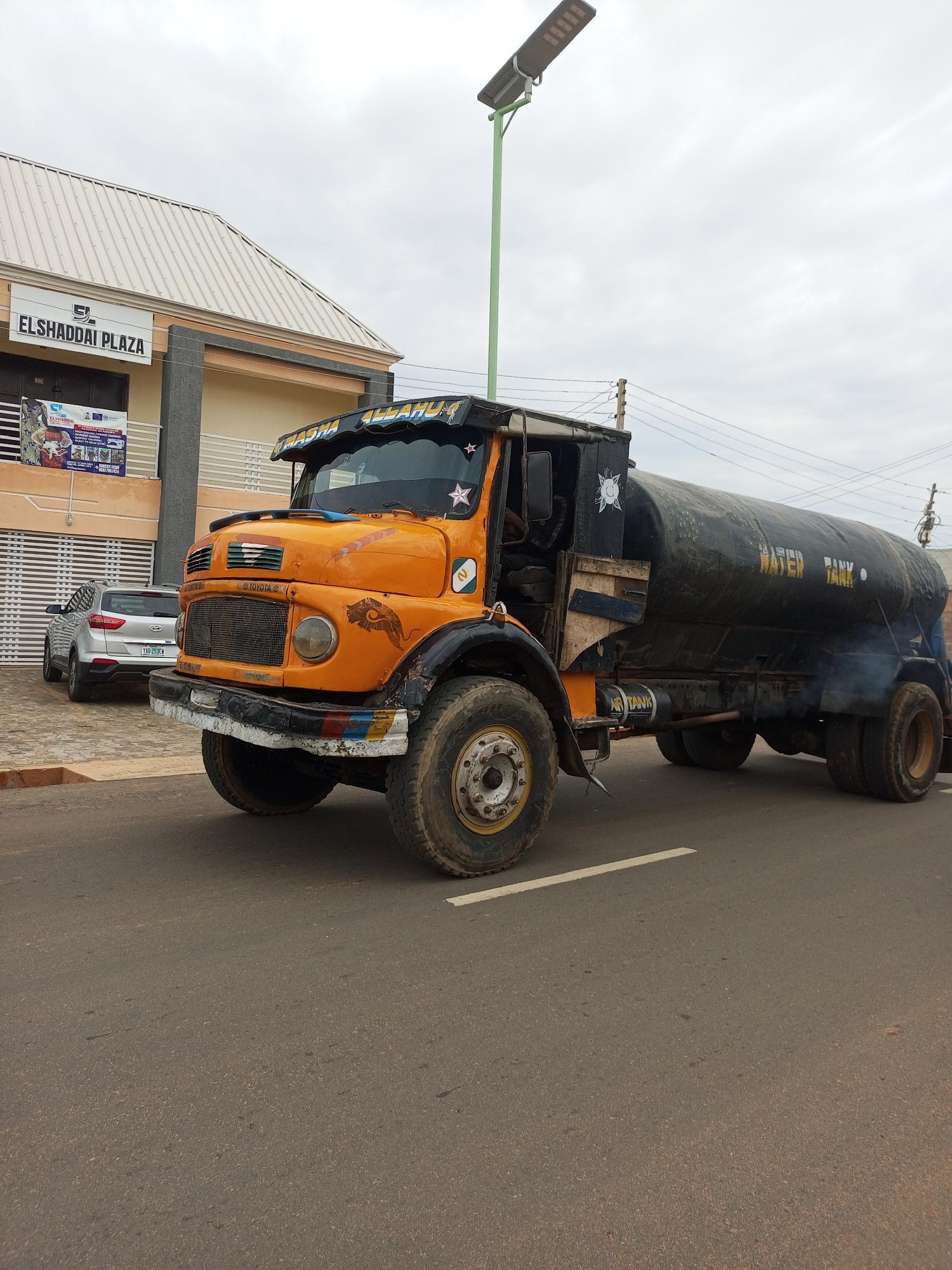 The image captures a street scene in Tempure, Nigeria, under an overcast sky. A large, orange water tanker truck is prominently parked on the right side of the asphalt road. The truck has "WATER TANK" printed in yellow on its side. To the left of the truck, a silver SUV is parked in front of a building. The building, labeled "ELSHADDAI PLAZA," is a light-colored, two-story structure with a light-colored metal roof and brown and grey accents. A solar-powered street lamp is visible on a green pole above the truck. The road has a white line marking its edge. Smoke emanates from the rear of the water tanker.