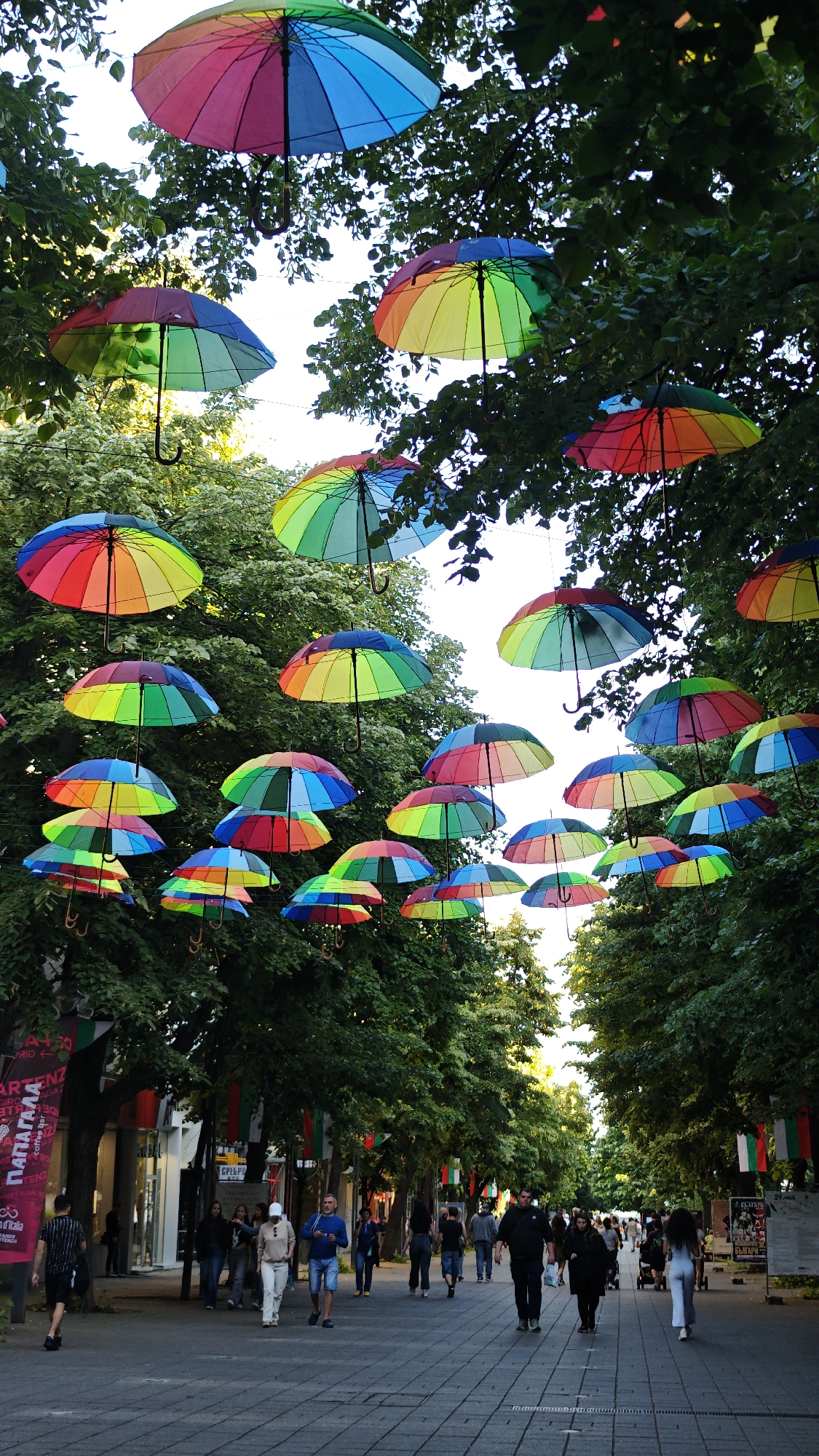 The image depicts a pedestrian street in Burgas, Bulgaria, characterized by vibrant rainbow-colored umbrellas suspended above the walkway between rows of tall, green trees. The street is paved with gray stone tiles, and pedestrians are walking along it. The pedestrians include a man in a black and white striped shirt, a couple walking together, and a woman in a white outfit. Bulgarian flags are hung along the sides of the street. Storefronts and advertisements are visible behind the trees on either side of the street. The sky is visible through the trees and between the umbrellas.