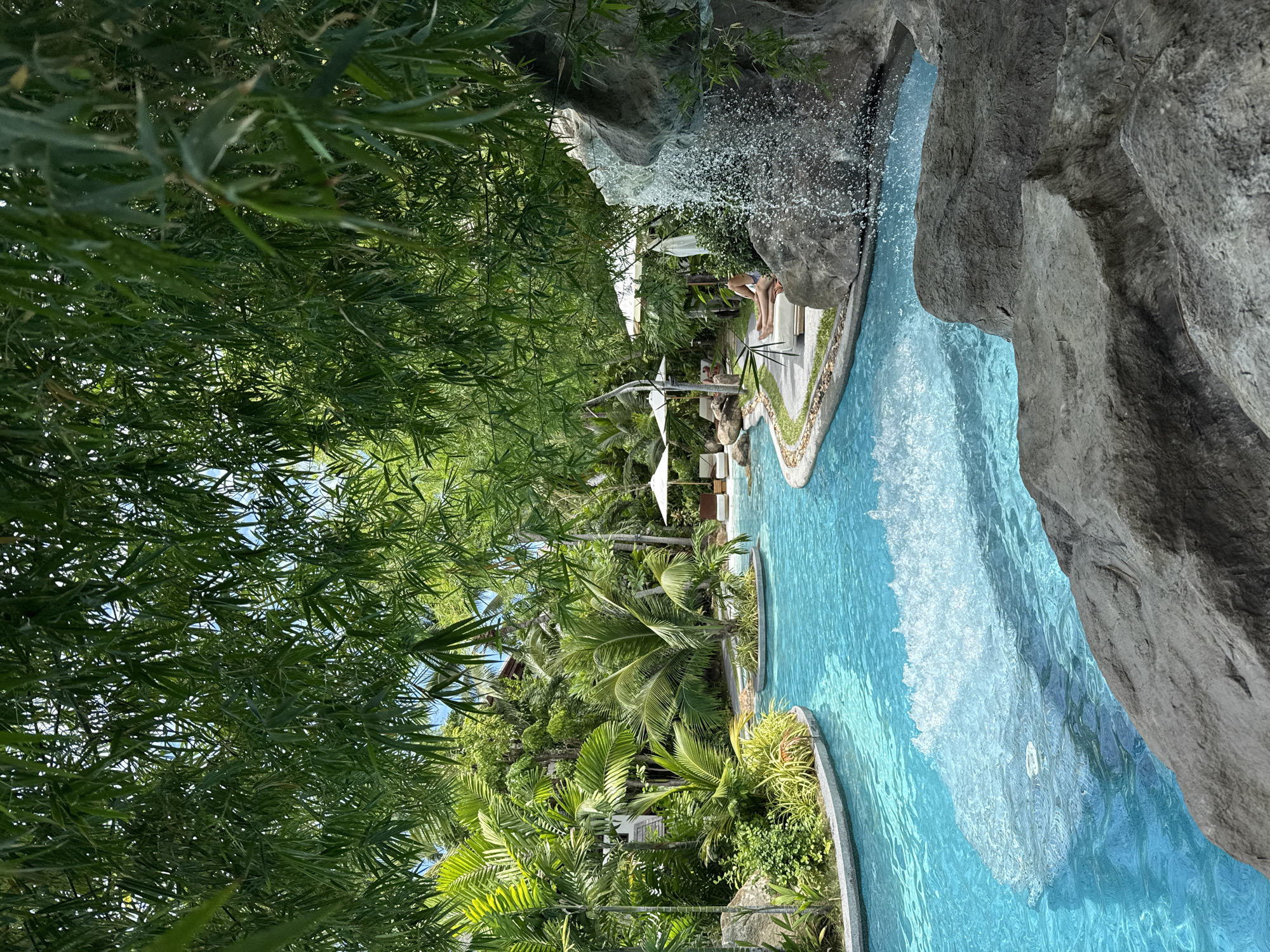 This image depicts a serene resort pool area in the Ko Samui District of Thailand. The pool, a vibrant turquoise blue, is bordered by lush tropical vegetation, including palm trees and dense greenery. A small waterfall cascades over large rocks on the right side of the frame, adding to the natural ambiance.
In the background, white umbrellas and lounge chairs suggest a relaxing atmosphere. A single individual is visible lying on a lounge chair near the waterfall, appearing to be sunbathing. The foreground is dominated by large, textured rocks, which create a sense of depth and frame the inviting pool. The overall impression is one of tranquility and luxurious escape amidst a tropical paradise.