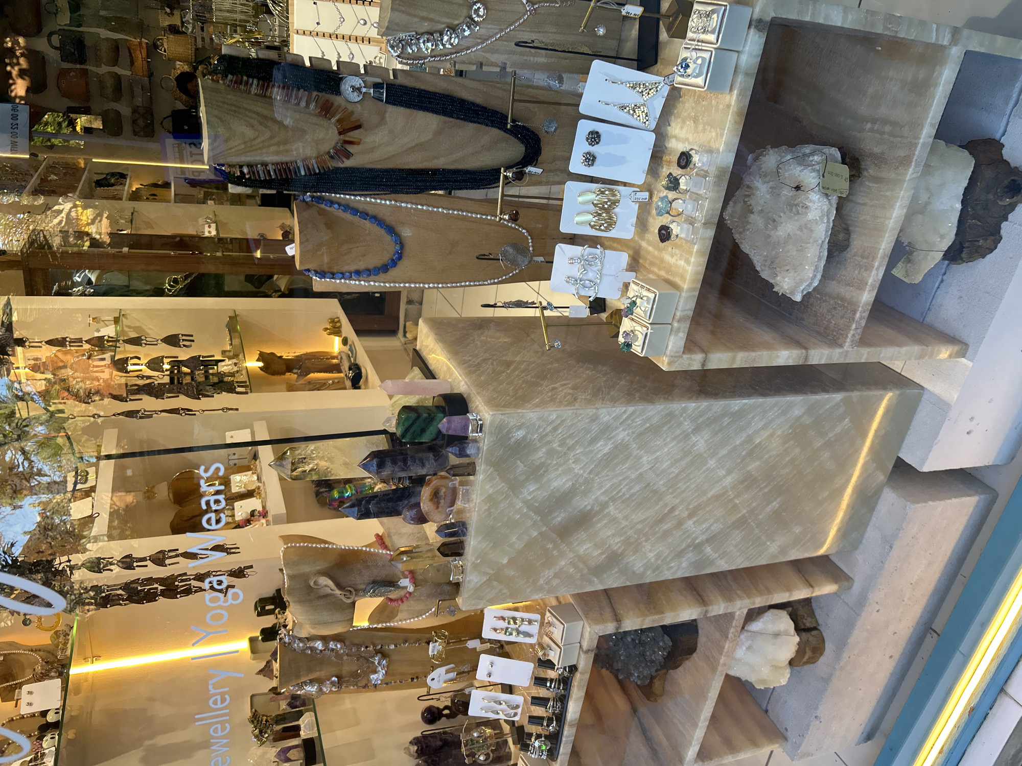 An indoor retail display window showcases a variety of handcrafted jewelry and decorative crystals. The window is organized with multiple shelves, some made of light-colored, translucent stone, and others of wood. On the shelves, there are necklaces with beads of various colors and sizes, displayed on wooden busts. Several pairs of earrings are presented on white cards, with rings and other small jewelry items also visible. Various crystal formations, including tall, dark points and smaller clusters in earthy tones, are strategically placed throughout the display. In the foreground, a large, rough-looking crystal chunk rests on a shelf. The background reveals more shelves with additional merchandise, including handbags, suggesting this is a boutique or specialty store. The overall lighting is warm, with a distinct yellow glow emanating from a light fixture on the left side of the window. The store name "Jewellery | Yoga Wears" is visible on the window in white lettering. The environment appears to be a shop interior, possibly in a tropical location, given the style of the merchandise. The specific location is indicated as Ubud District, Indonesia. No people are present in the immediate view of the display.