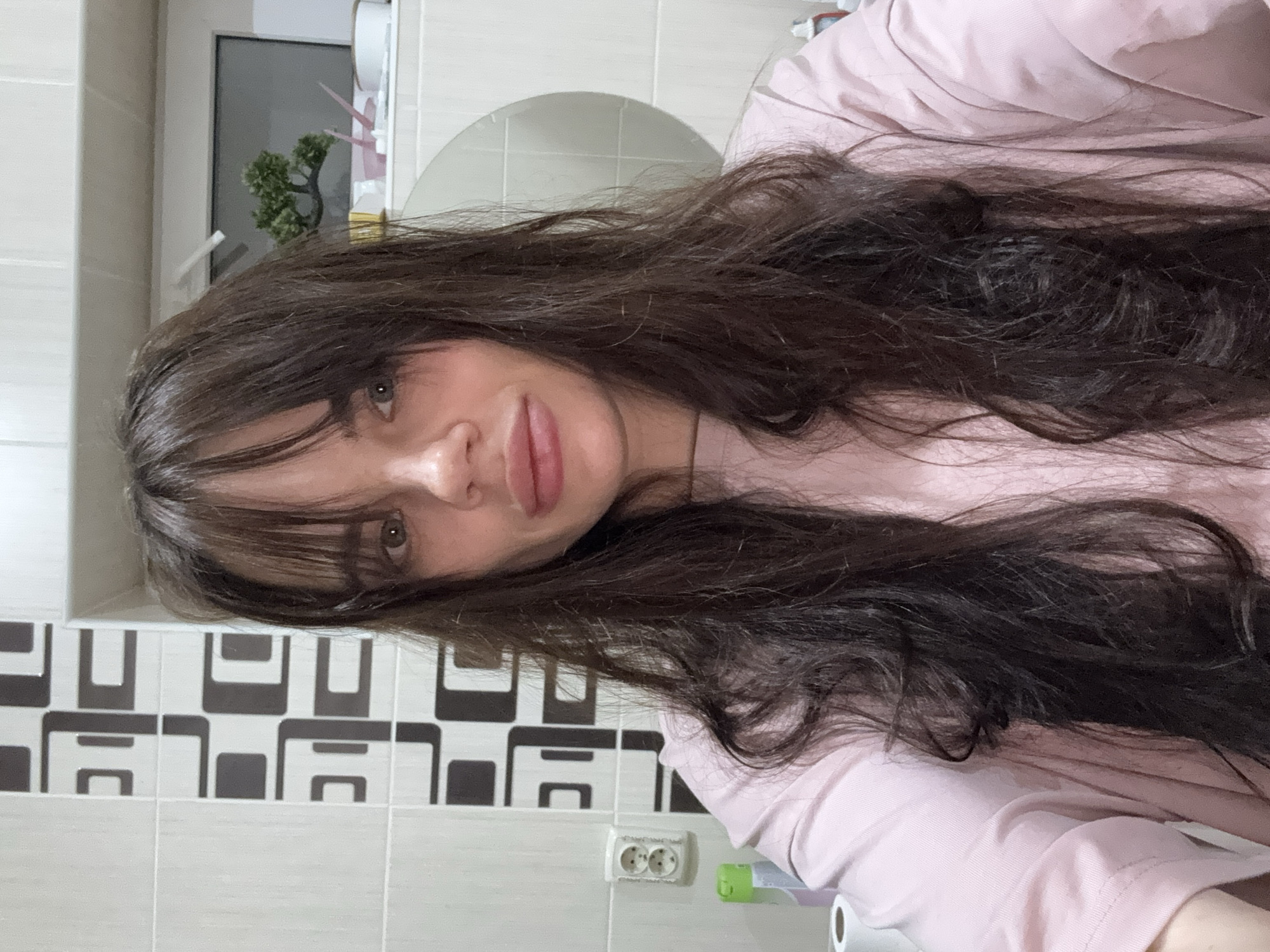 A close-up portrait shows a young woman, likely in her twenties, from the chest up. She has long, dark brown hair with bangs framing her face, which falls loosely over her shoulders and chest. Her eyes, which appear to be a dark hazel or brown, are open and look directly at the viewer. Her lips are full and slightly parted. She wears a plain, light pink short-sleeved top. The image is oriented vertically, making it appear as if she is lying on her side or leaning against a surface, with her head tilted.
The background suggests an indoor setting, specifically a bathroom, within the location context of Rostov-on-Don, Russia. Behind her, a wall is visible, partly tiled with a beige-grey pattern, and partly with a distinctive black and white geometric pattern of interlocking squares and rectangles. To her lower right, a white electrical outlet with two circular sockets is affixed to the tiled wall. Other elements in the background include a small, green artificial plant resembling a bonsai, a white toilet paper roll, and a green object that looks like a toothbrush holder or a toothbrush. The lighting appears to be indoor ambient light, and no specific time of day or weather conditions can be discerned. Her expression is neutral and calm, suggesting a casual selfie or portrait. There is no visible text in the image.