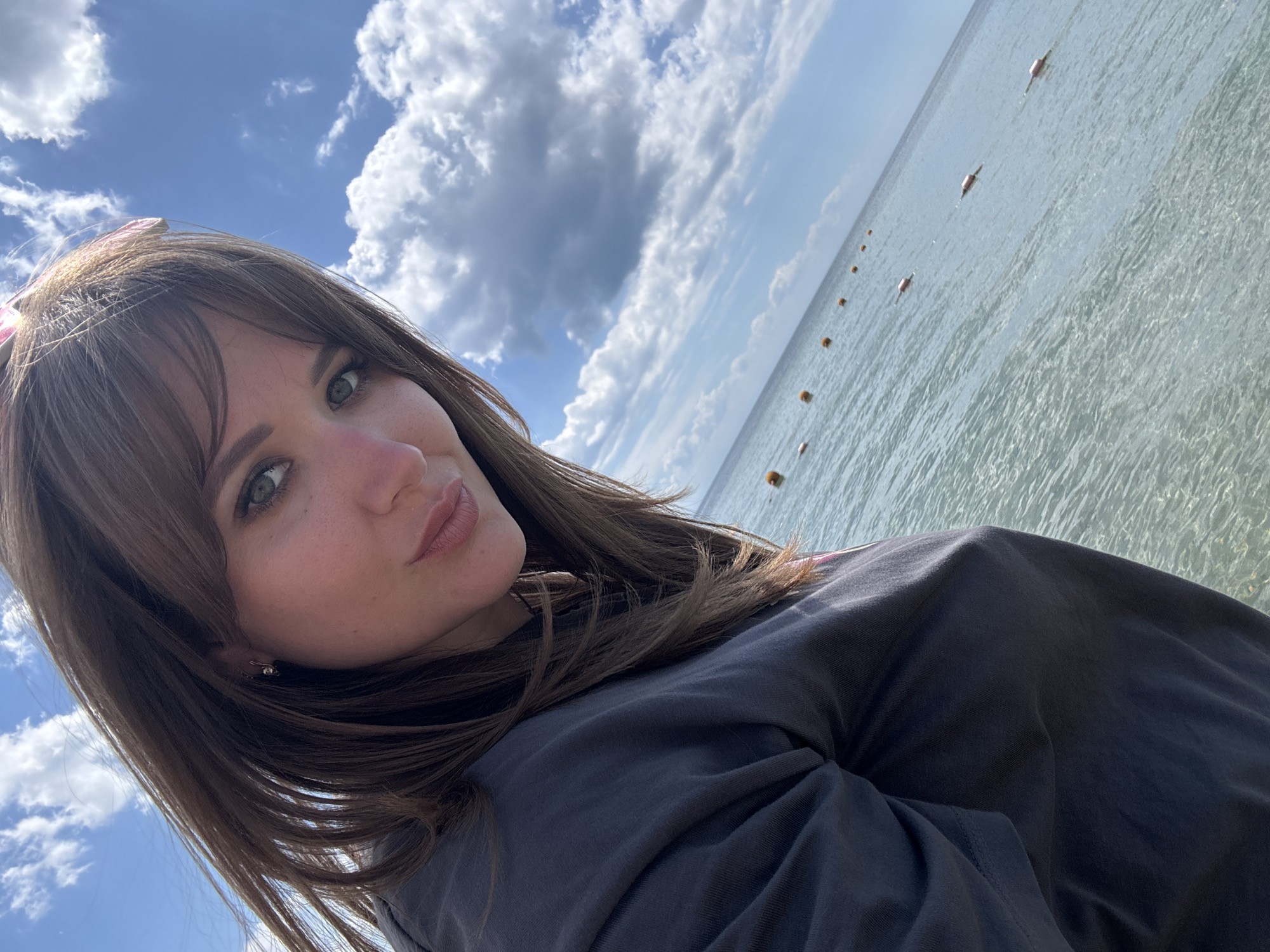 A woman with brown hair and green eyes takes a selfie outdoors. She's wearing a dark gray t-shirt. Behind her is a body of water with a line of orange buoys stretching into the distance. The sky is blue with fluffy white clouds. The location is Yevpatoriya, Ukraine.