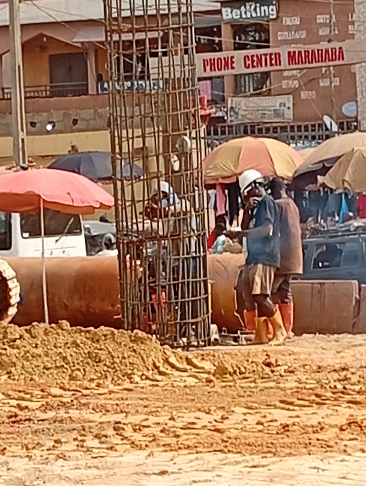 The image shows a construction site in Ado, Nigeria, based on the "PHONE CENTER MARARABA" and "BetKing" signs in the background. A vertical rebar cage stands prominently in the foreground, with workers nearby. One worker is wearing a white hard hat, dark shirt, and light-colored shorts and rubber boots. A second worker in an orange shirt, brown trousers and red boots stands behind him. Large orange pipes are also present. There are umbrellas and vehicles in the background, suggesting a busy area, and piles of dirt occupy the foreground.