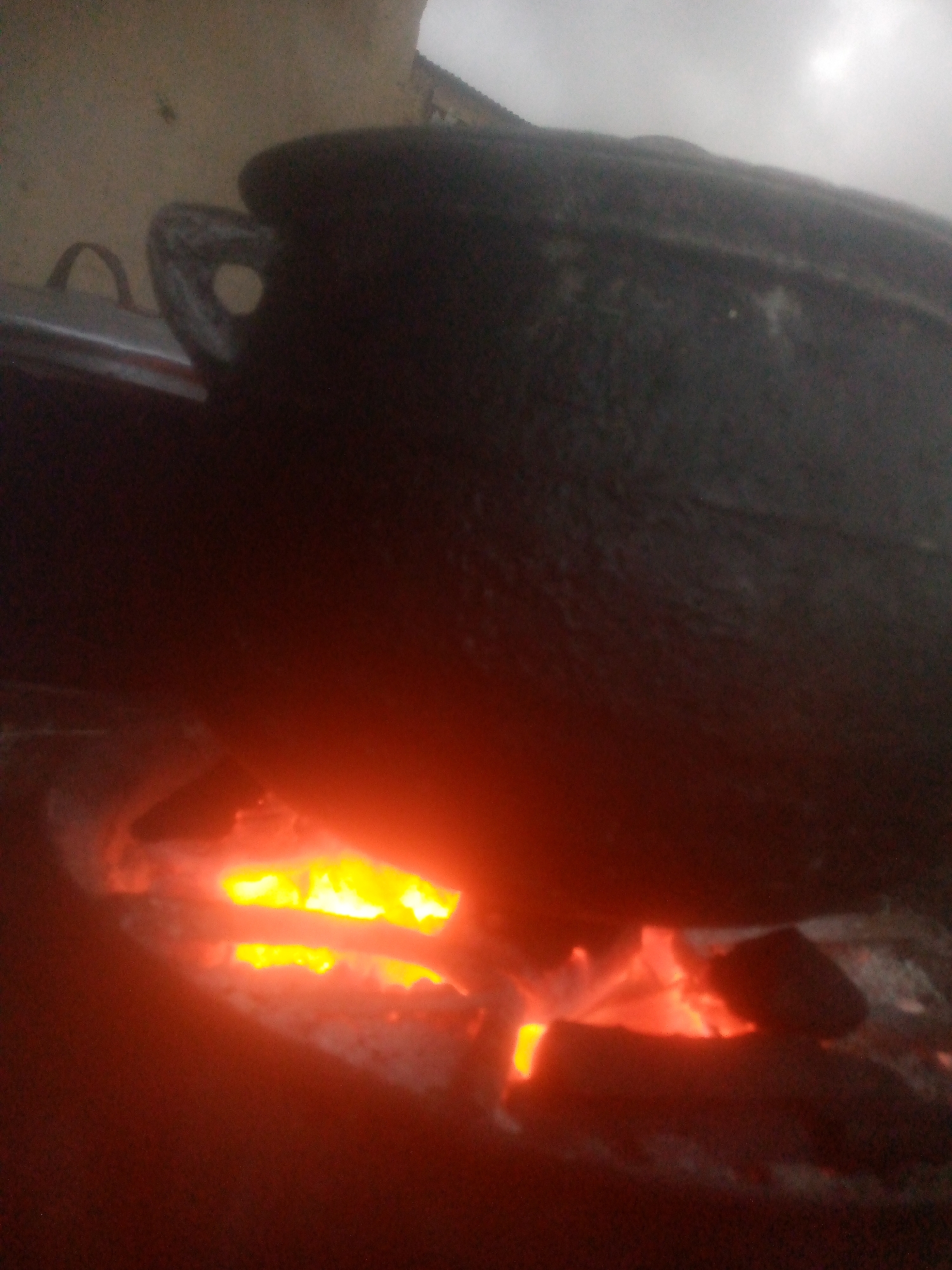 A black cast iron pot sits on a wood fire. The fire glows orange and yellow, illuminating the bottom of the pot and the surrounding area. The pot has a handle with a hole in it, visible on the left side. The background is dark and out of focus, suggesting an indoor setting. The location is unknown.