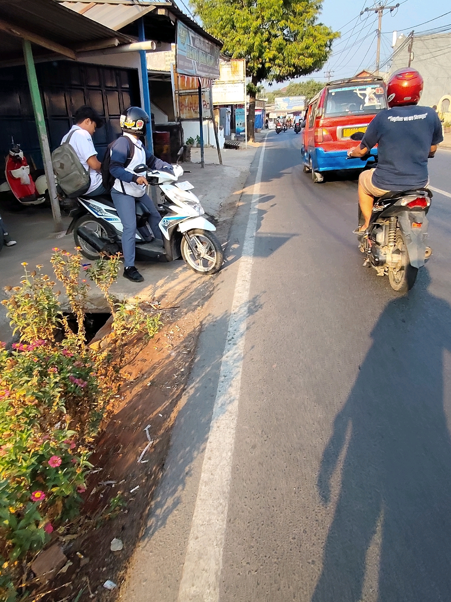 The image captures a street scene in Purwakarta, Indonesia, under a clear sky with the sun casting long shadows. In the foreground, a person on a white scooter, wearing a white shirt and dark pants, is parked by the side of the road. To the left of this scooter, another scooter, red and white, is parked and a person wearing a white shirt and a backpack is seated on it. A red and blue van is seen further down the road, moving away from the viewer. Behind the van, a person on a black motorcycle is riding away from the viewer, wearing a red helmet and a dark blue shirt. The environment includes a sidewalk with some vegetation, a drain, and various structures along the roadside, suggesting a commercial or residential area. The overall atmosphere is that of a typical day in a bustling Indonesian town.