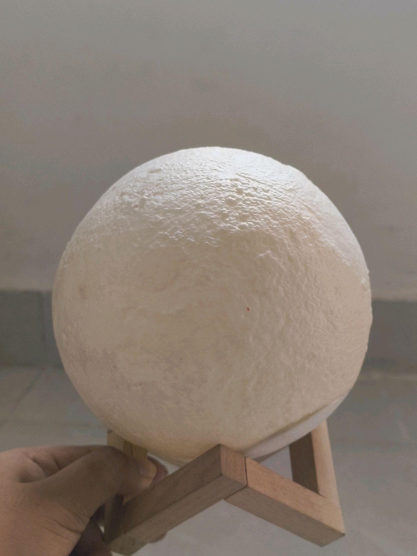 A close-up, eye-level shot shows a person's hand supporting a textured, spherical object, possibly a lamp or decorative item resembling a moon, from below. The object is white with a matte finish and exhibits a cratered or bumpy surface. It rests on a light-colored wooden stand with two angled legs that form a V-shape at the bottom. The background is a plain, neutral-toned wall with a subtle texture, and a faint horizontal line appears near the top of the frame, suggesting a separation between wall sections or a shelf. The lighting is soft, creating subtle shadows on the object and its stand. The image was taken indoors. The location of this scene in the city of Mohali, India, is not discernible from the visual information provided.
