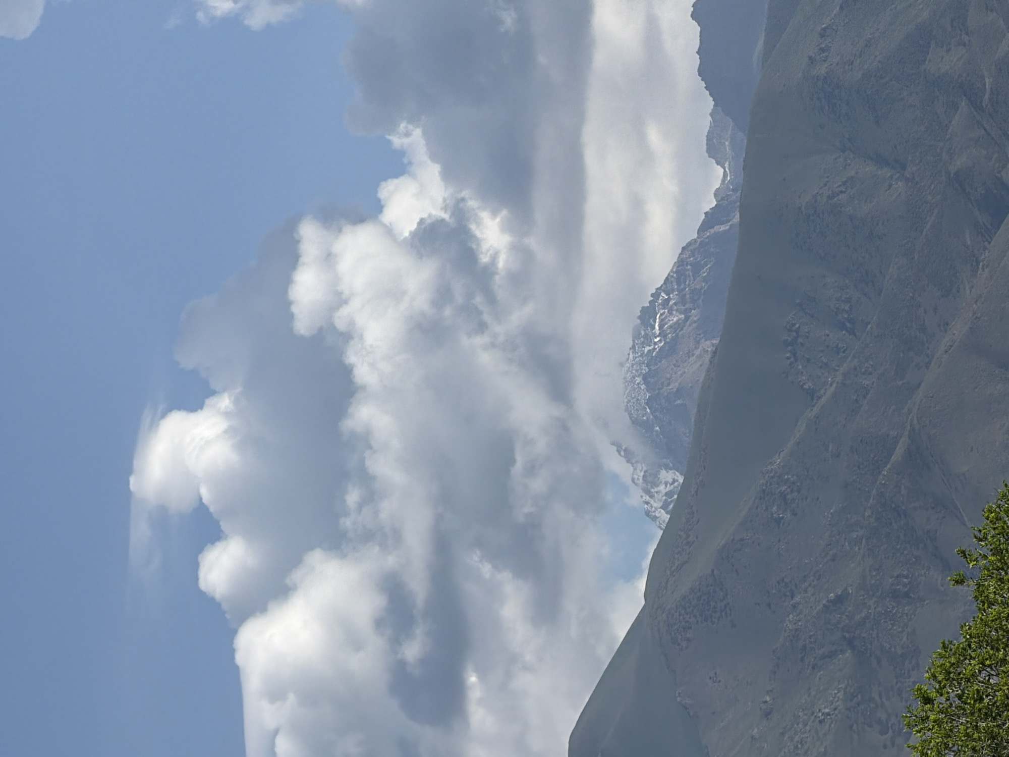 The image displays a mountainous landscape under a partly cloudy sky. The foreground features green foliage, likely from trees. Behind the foliage are rolling, green hills that recede into the distance. The midground is dominated by the slopes of these hills, which show varying textures and contours, some appearing rocky and barren. In the background, the highest peaks are visible, capped with snow and jagged rock formations. The sky is a mix of blue and white clouds, with some clouds obscuring parts of the mountain peaks. The lighting suggests it is daytime, with the sun likely positioned behind the clouds or to the side, casting diffused light on the scene. There are no people or man-made objects clearly visible in the frame.