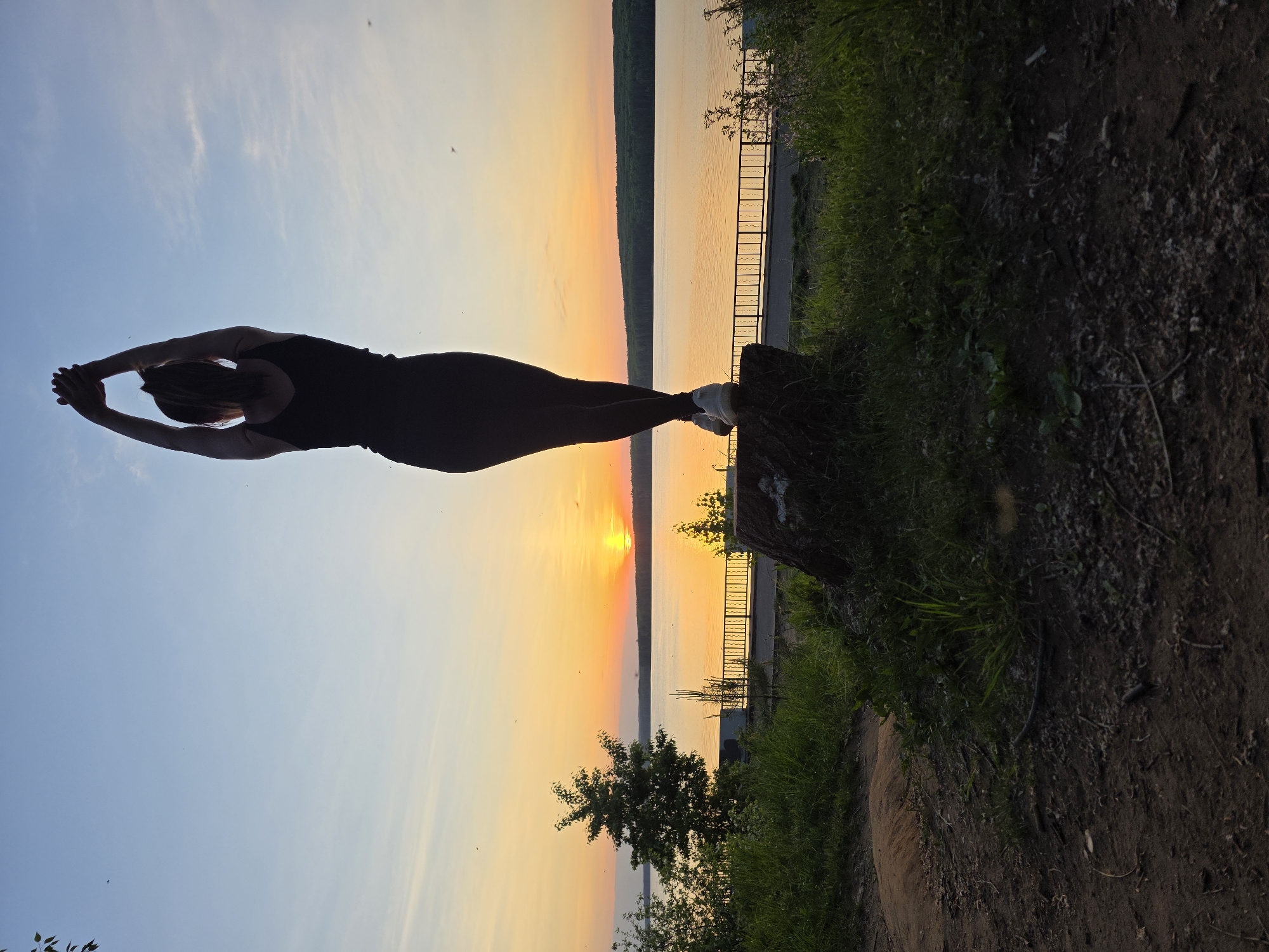 A person is standing on a tree stump in Chaikovsky, Russia, with a body stretched out horizontally towards a sunset over a body of water. The individual is wearing dark clothing and white shoes, with arms outstretched in front. The background features a lake, a fence, trees, and a sunset with a yellow to orange gradient. The ground around the stump is covered in grass and dirt.