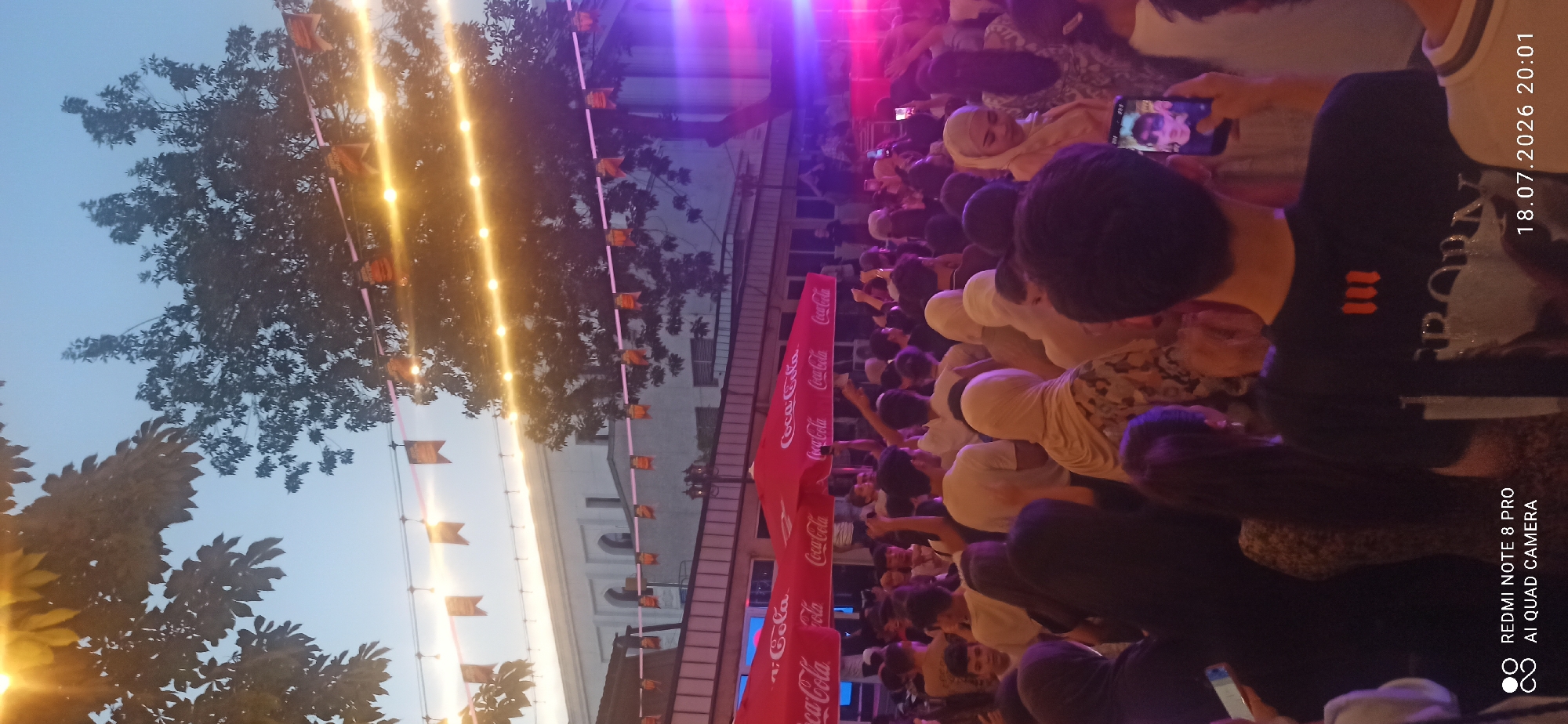 An outdoor gathering takes place at dusk in Dushanbe, Tajikistan. The scene is illuminated by string lights and vibrant stage lighting, casting a colorful glow. In the foreground, a crowd of people, many wearing hijabs, are visible from behind. Some are holding up their phones, suggesting they are capturing the event. In the midground, red Coca-Cola branded umbrellas provide shade. Behind them, a building with arched windows is partially visible. The overall atmosphere suggests a lively social event.