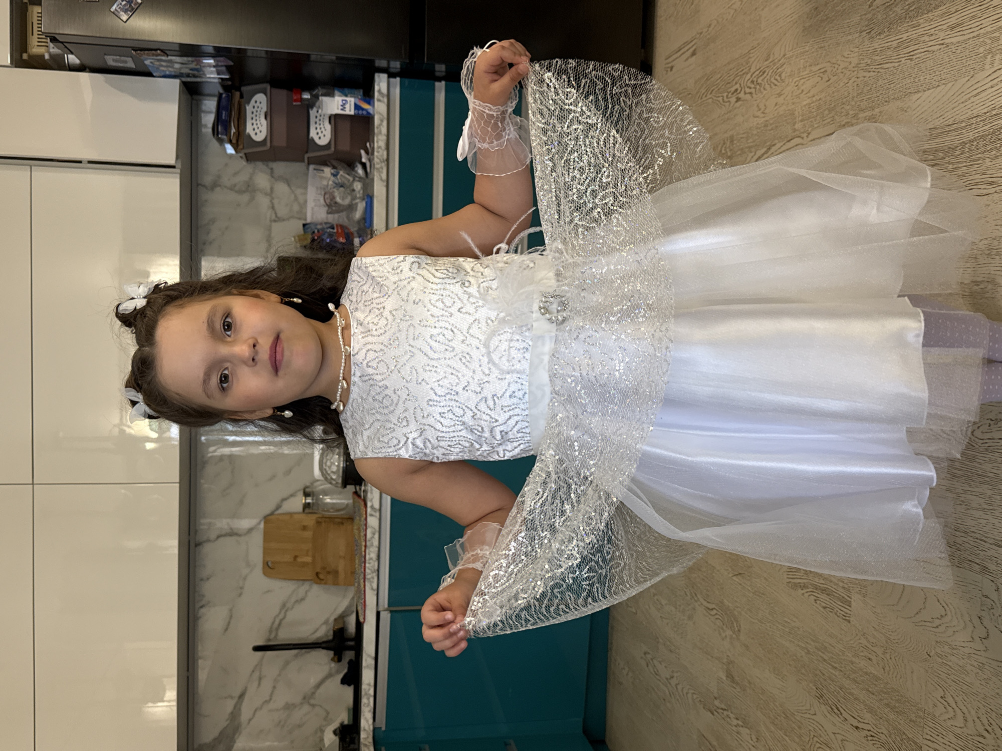 A girl is indoors, dressed in a white formal dress. She is holding a part of the dress's net overlay up with her hands. The location appears to be a kitchen in a residential setting, with white cabinets and a marble-patterned backsplash visible behind her. A refrigerator is also visible. The city of Izhevsk, Russia, cannot be confirmed or denied based on this image.