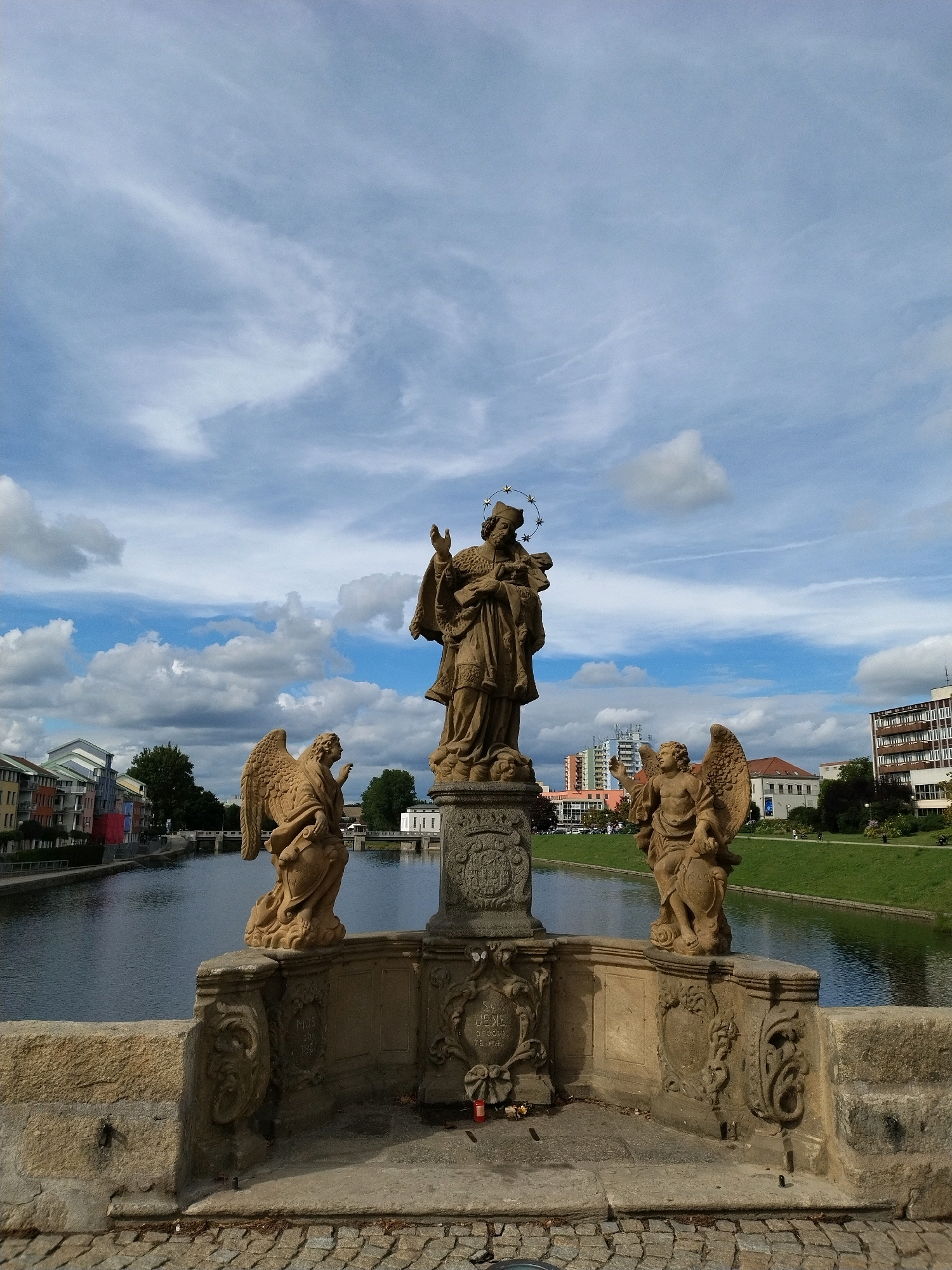 The image captures a view of the Old Bridge in Písek, Czechia, dominated by a central statue of Saint John of Nepomuk and flanked by two angel statues. The statues are made of stone and are positioned on a bridge with a river flowing below. The bridge itself has stone balustrades with intricate carvings. In the background, there are buildings lining the riverbank, suggesting an urban setting. The sky is partly cloudy, with patches of blue sky visible between white and grey clouds. The overall impression is one of a historical monument in a city environment.
