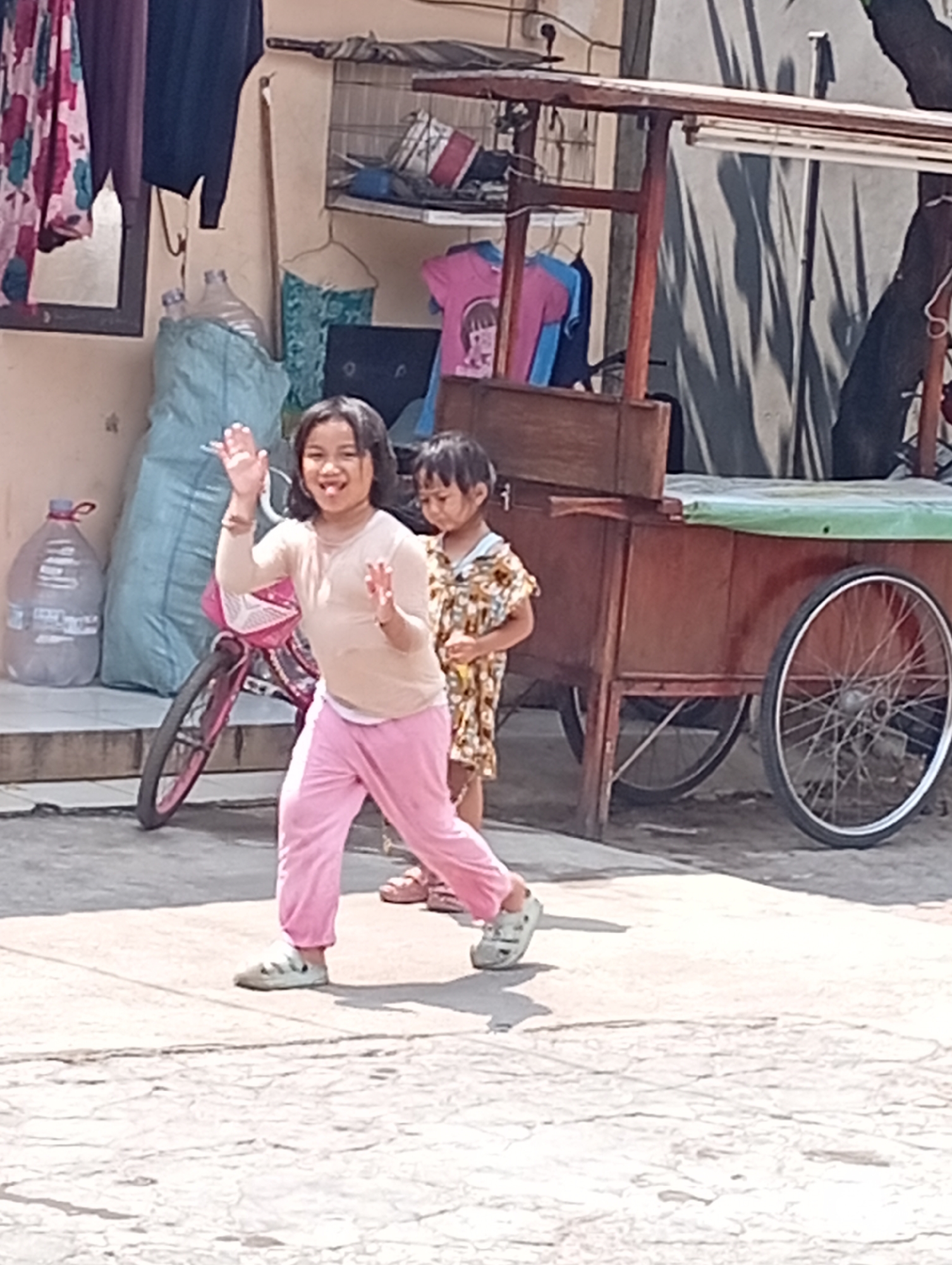 In a street scene in Bekasi, Indonesia, two young girls are interacting in front of a wooden cart. The girl in the foreground, wearing a light beige long-sleeved shirt and pink pants, smiles widely and waves both hands, her tongue visible. She is wearing light-colored clogs. Behind her, a younger girl in a patterned yellow and brown dress stands partially obscured. To the left, a large plastic water bottle and a bicycle with a pink basket are visible. The background features a building with hanging clothes and a cage. The wooden cart, with its dark wood frame and a green tarp covering its top, has a large spoked wheel on its right side, and a smaller wheel is partially visible underneath. The ground is paved with cracked concrete.