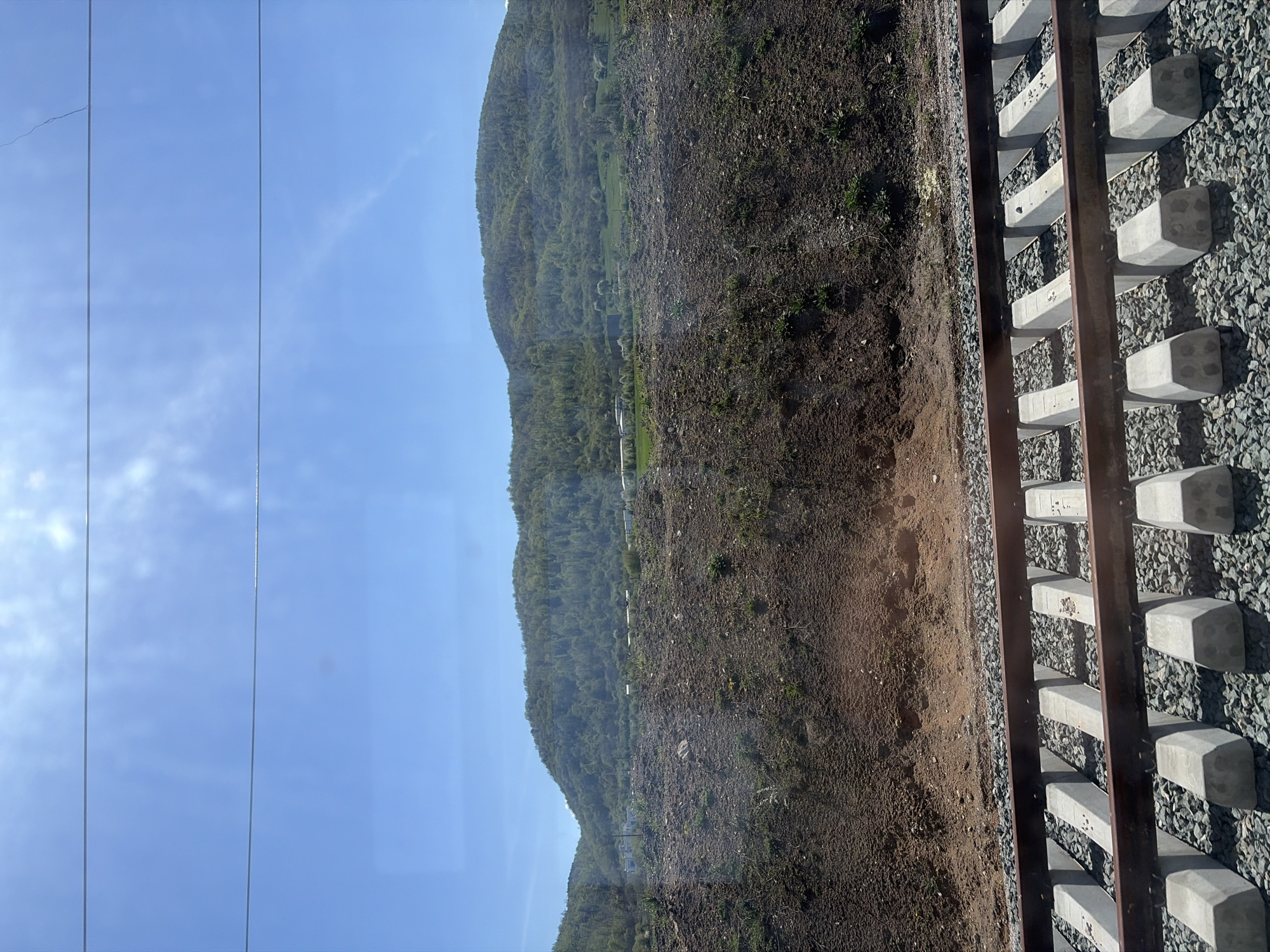 The image shows a view from a train window. On the left is a clear blue sky with power lines. In the center is a hillside covered with sparse vegetation and brown soil. On the right are railroad tracks with concrete sleepers and gravel. The location appears to be rural. The scene may be near Stambolovo, Bulgaria, given the prompt. No people or actions are visible.