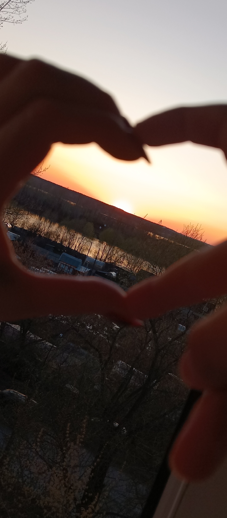 A person is holding their hands in a heart shape, framing a view of a river and sunset. The background shows a river with trees lining the banks, buildings, and industrial structures in the distance. The sky is orange and yellow from the setting sun. The foreground shows trees and buildings. The location is unknown.