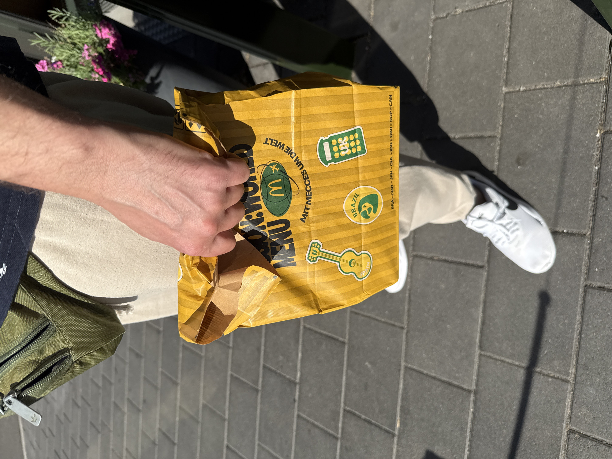 A person is holding a yellow McDonald's "McRworld" menu bag, which features various country-specific icons and the phrase "Mit MecceS um die Welt" (with McDonald's around the world). The bag has images of a guitar with "Spain" written below it, a toucan with "Brazil" written on it, and what appears to be a calculator. The McDonald's logo is also visible on the bag. The person is wearing beige pants and white sneakers, and a green fanny pack is visible around their waist. The bag is held in front of a paved walkway, indicating an outdoor setting in a city. The shadow of the person and the bag are cast on the ground, suggesting it is a sunny day. The general environment suggests a commercial area, possibly near a McDonald's restaurant. The image does not contain any specific visual cues that definitively place it in Werther, Germany.