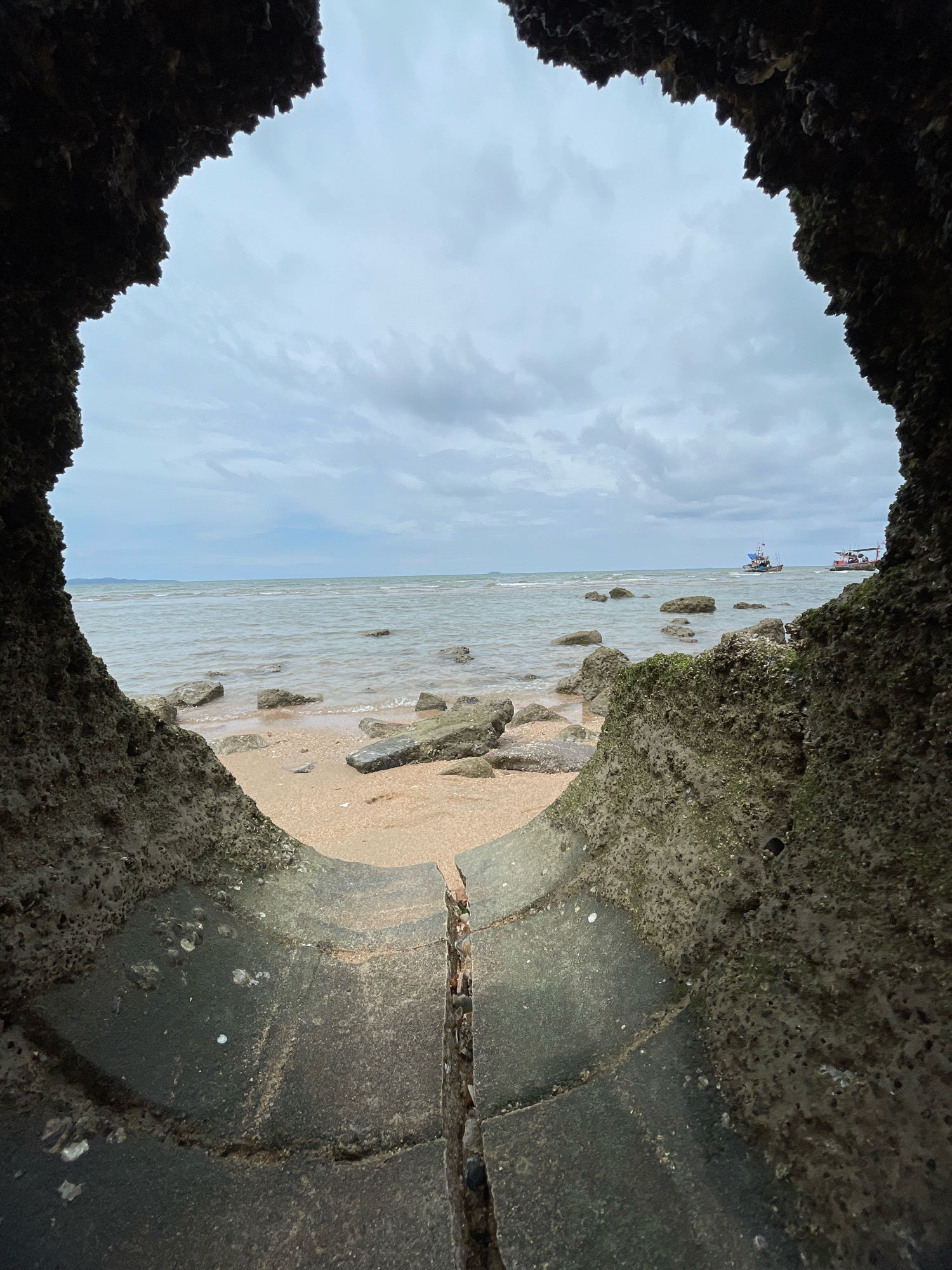 The image shows a view of the sea from within a large concrete pipe opening. The pipe's interior is dark and rough, with barnacles and other marine growth visible on its surface. The opening frames a beach with sand and scattered rocks, leading to the calm sea. Two small fishing boats are visible in the distance on the water, under a cloudy sky. The scene appears to be a coastal area in Ban Na Chom Thian, Thailand.