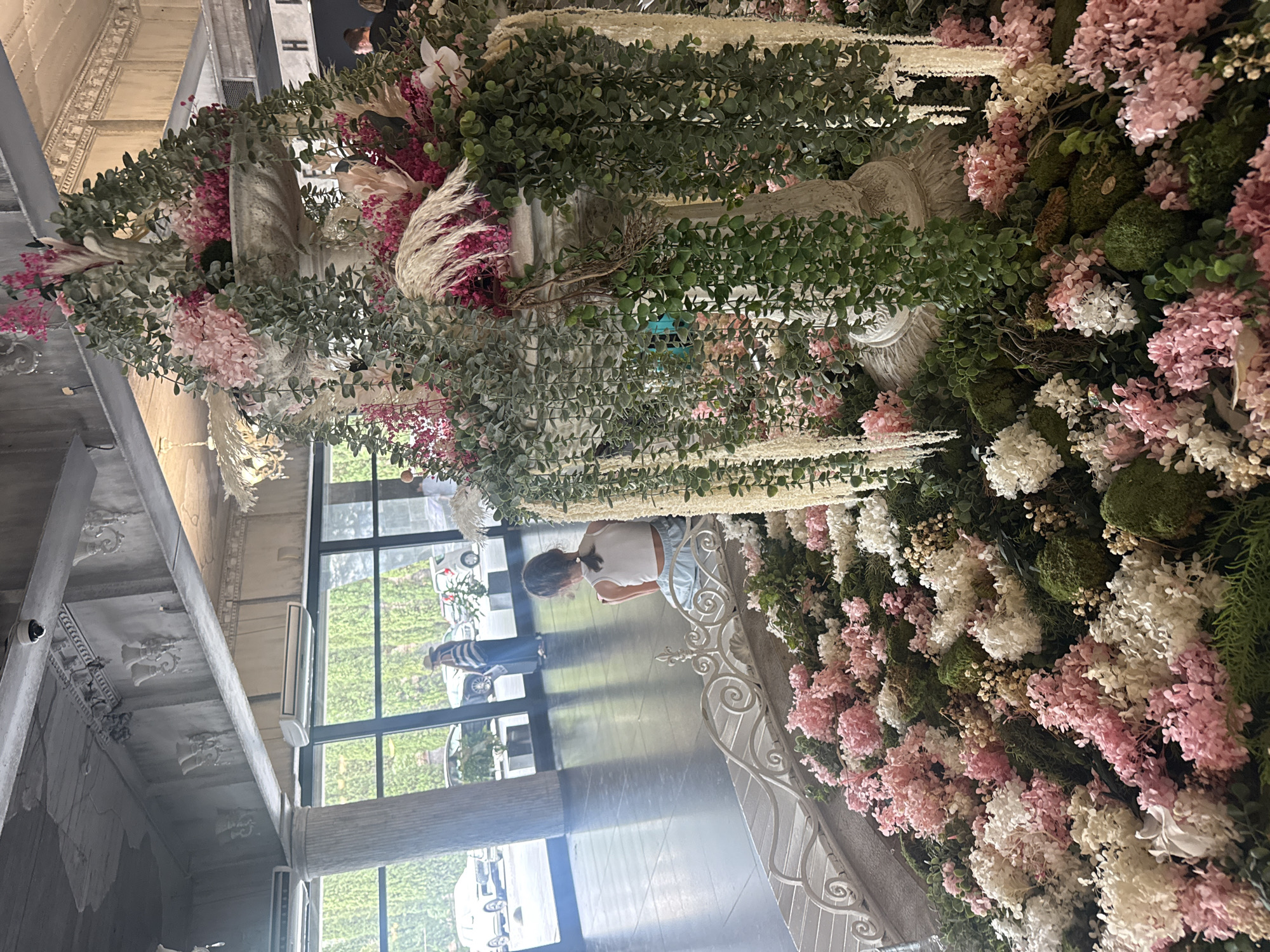 A young girl with her back to the camera is sitting on a decorative white railing. She is wearing a white tank top and light blue shorts. In front of her, a large tiered fountain is elaborately decorated with pink and white flowers, moss, and cascading greenery. The fountain is situated in what appears to be a hotel lobby or event space, with large windows in the background revealing a street scene with cars and pedestrians. The ceiling features ornate moldings and what looks like a security camera. The flooring is a dark, reflective tile.