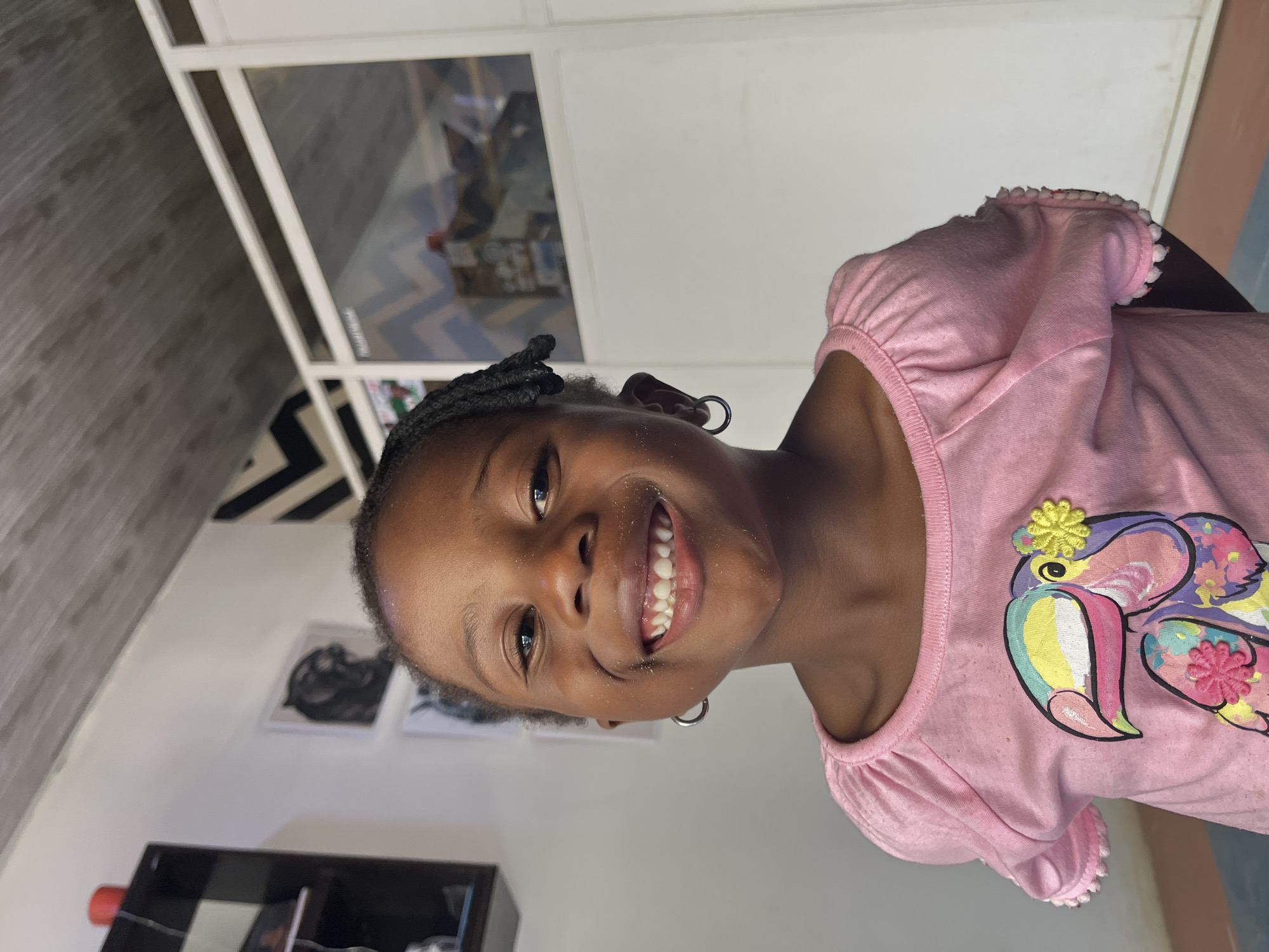 A young girl with braided hair and silver hoop earrings smiles at the camera. She wears a pink short-sleeved shirt with a cartoon toucan design on the front. She has a dusting of what appears to be sand around her mouth and nose. The setting appears to be indoors, possibly a home in Jos, Nigeria, given the mention of the city. The background includes a white wall with framed pictures, a black shelving unit, and a window.