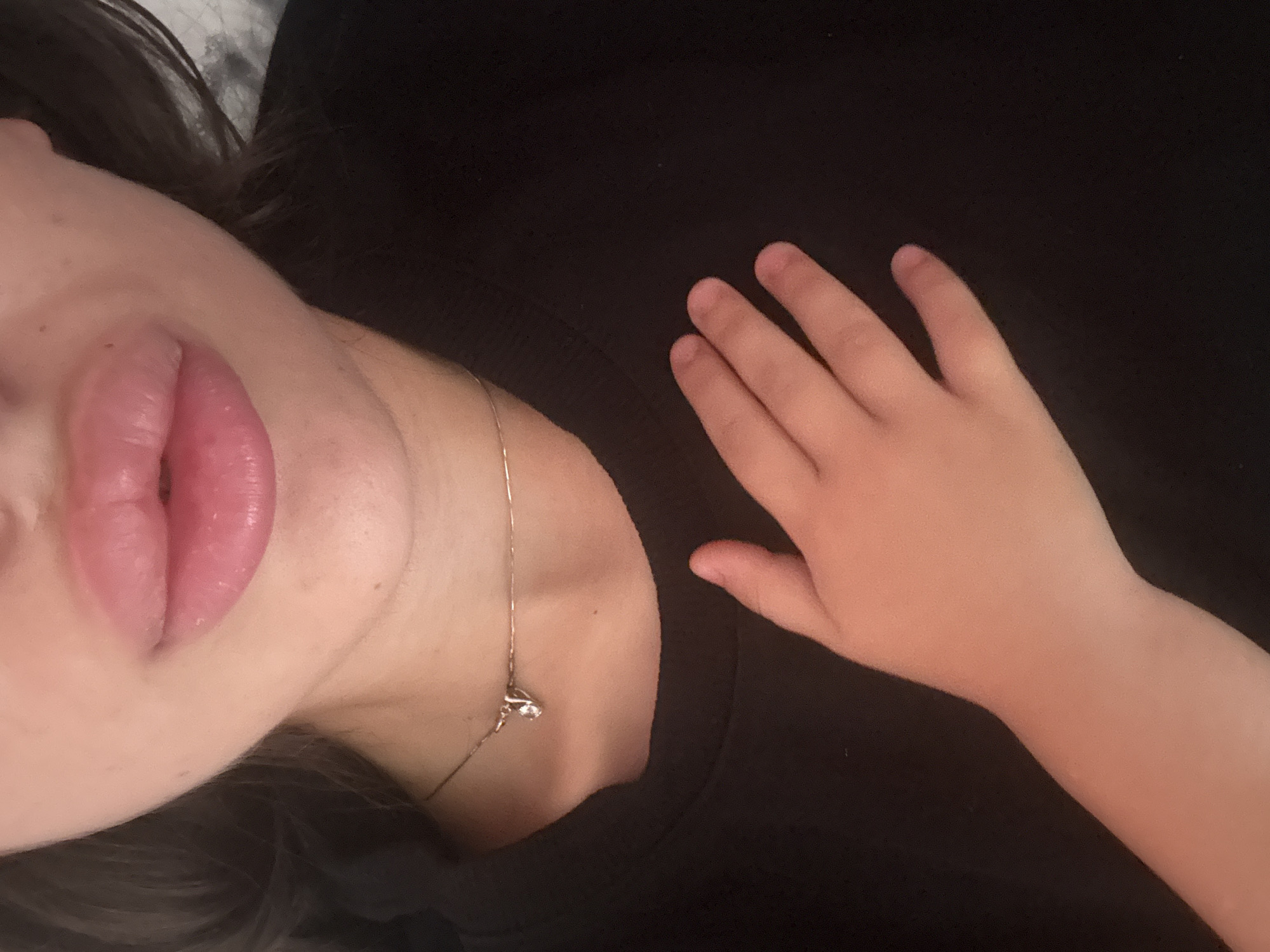 The image shows a close-up of a person's lips and upper chest, with a child's hand placed over the chest. The person's lips are noticeably plump and pink. They are wearing a dark-colored sweatshirt and a thin necklace with a small pendant. The child's hand appears to be resting on the sweatshirt. The background is dark and out of focus. There is no discernible information about the location within the city of Basarabeasca, Moldova.