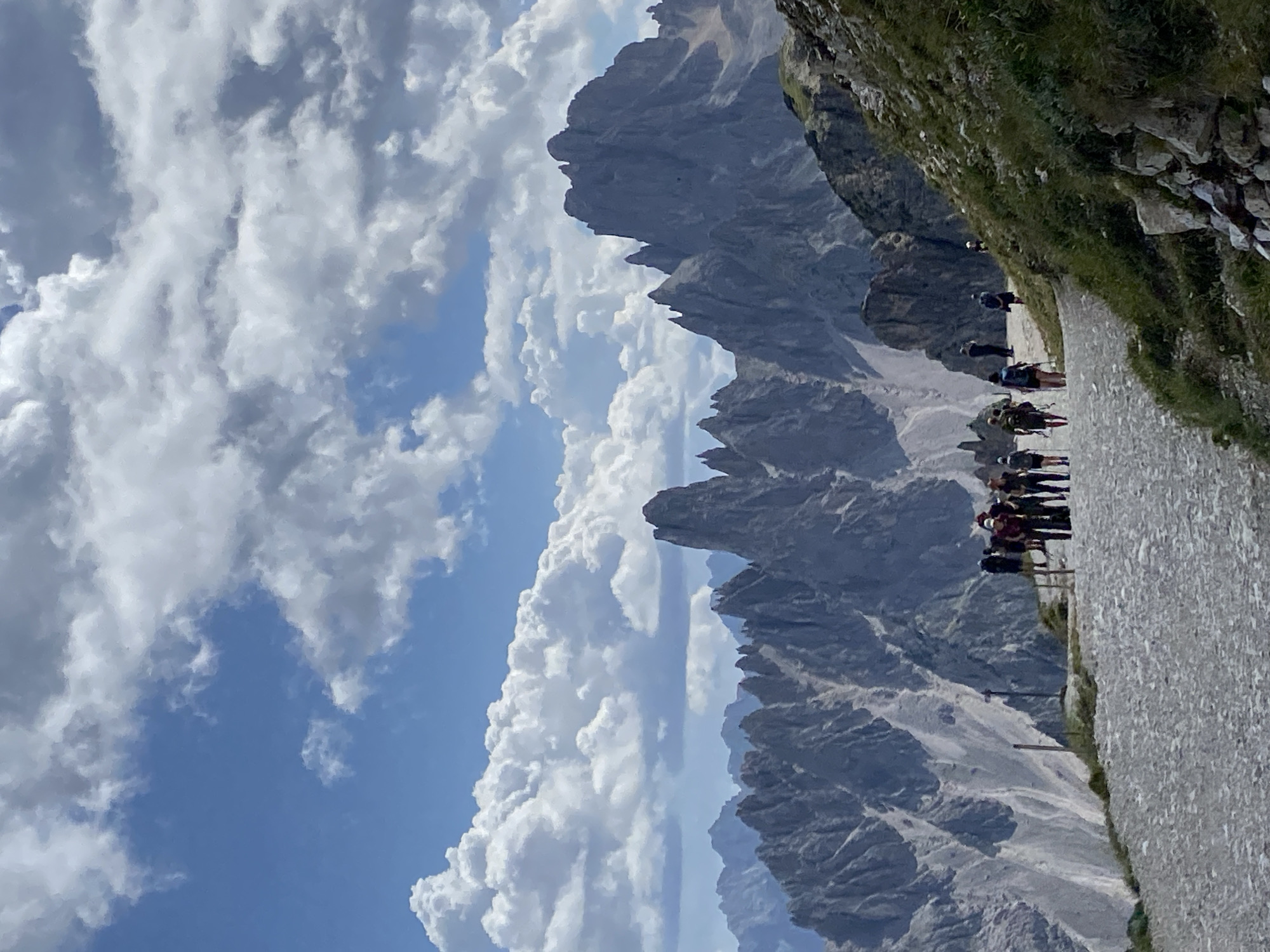 This image depicts a scenic mountain landscape under a partly cloudy sky, likely in the vicinity of Auronzo di Cadore, Italy, renowned for its dramatic Dolomite peaks. The foreground features a wide, gravel path leading uphill, populated by a group of approximately ten hikers, some of whom are using trekking poles. The hikers are seen walking in the same direction, suggesting a shared activity or route.
Beyond the path, the landscape unfolds into a rugged mountain range characterized by sharp, jagged peaks of gray rock. The mountains exhibit steep inclines and deep ravines, indicative of a high-altitude environment. The sky is a vibrant blue, punctuated by large, fluffy white clouds that cast shadows on the mountains, adding depth and texture to the scene. The overall impression is one of a popular hiking destination, where natural beauty and outdoor recreation converge.