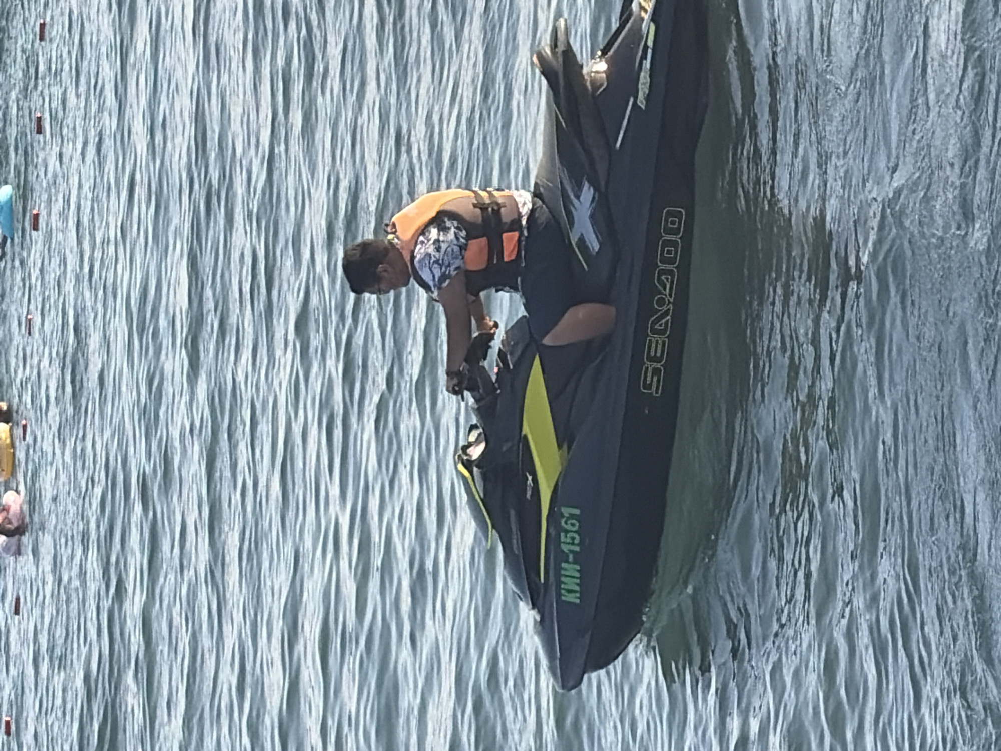 An adult male wearing a life vest over a patterned shirt and dark shorts is riding a black and yellow jet ski on choppy water. The jet ski has the registration number "КИН-1561" and "SEA DOO" written on it. The background shows a hazy view of buoys and people in the water. The scene appears to be a recreational area.