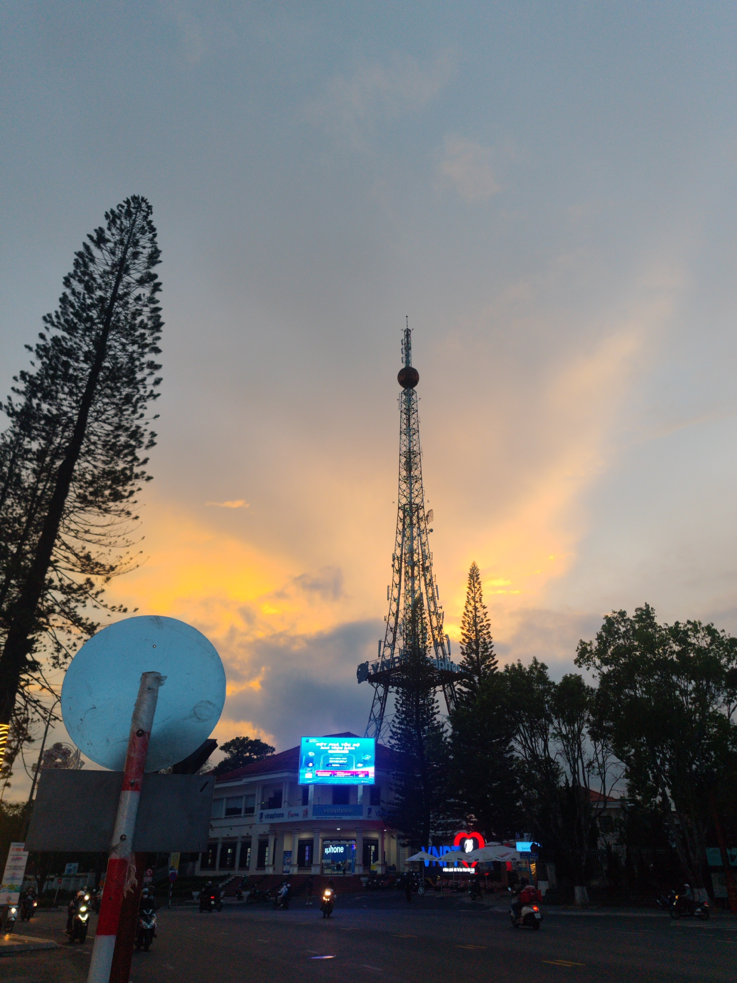 A low-angle view of a city street scene in Da Lat, Vietnam, at dusk. The sky is streaked with orange and gray clouds. A tall communication tower dominates the skyline, situated behind a building with a large blue digital display screen. The building is identified as a "Vinaphone" establishment.
Several trees, including a tall, slender pine, frame the tower and building. A round, faded blue sign is positioned on a red and white striped pole in the lower left foreground. The street is dark, with headlights visible from motorbikes traveling in both directions. A small number of people can be seen near the Vinaphone building.