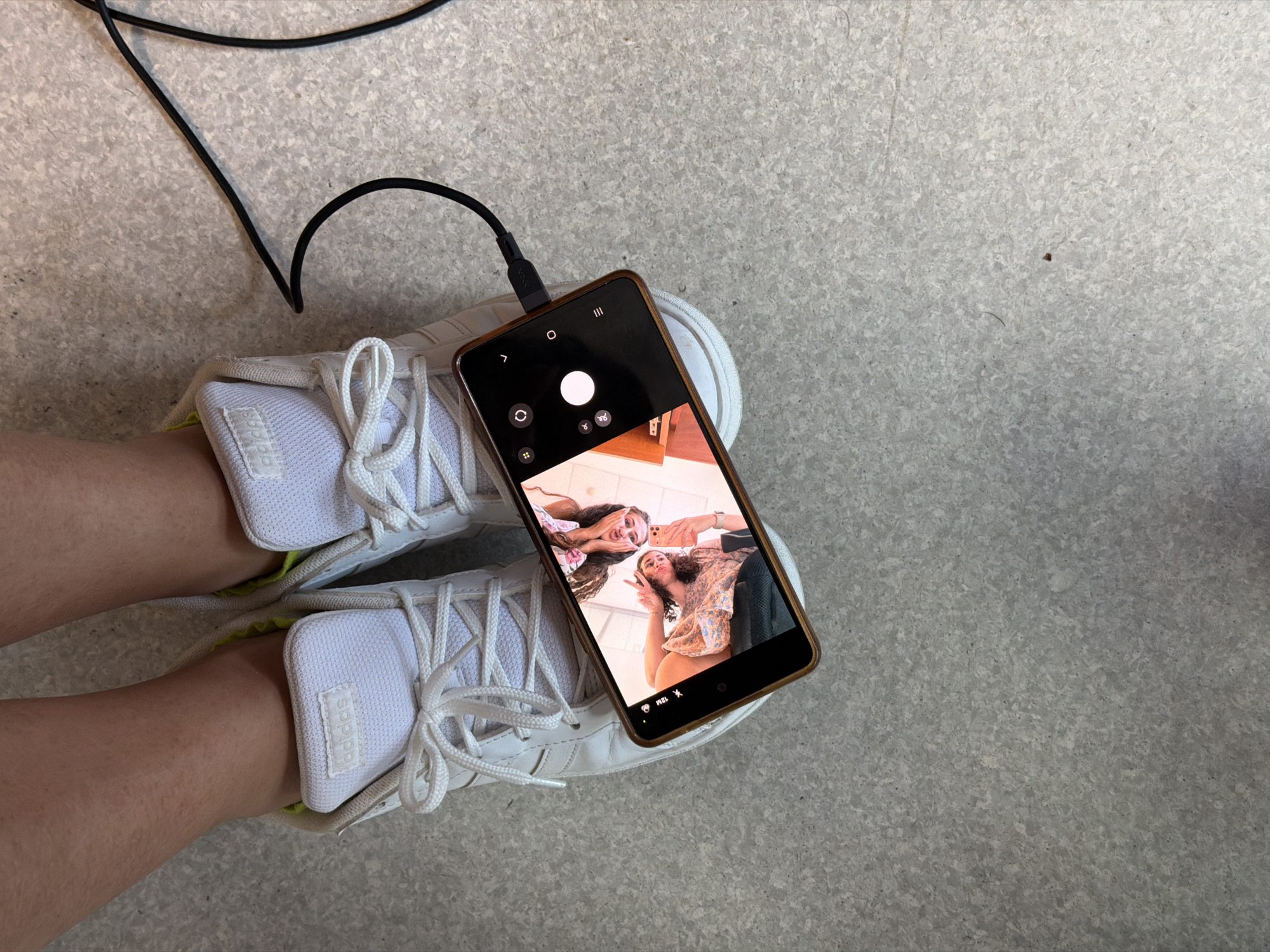 An overhead shot shows two pairs of white Adidas sneakers on a speckled gray surface. A smartphone is placed horizontally across the shoes, displaying a selfie of two young women. The woman on the left has long brown hair and is wearing a floral top. The woman on the right has curly brown hair and is wearing a patterned dress. She is holding the phone and making a peace sign. A black charging cable is connected to the phone and extends out of the frame to the right. The scene is devoid of any specific city or location indicators.