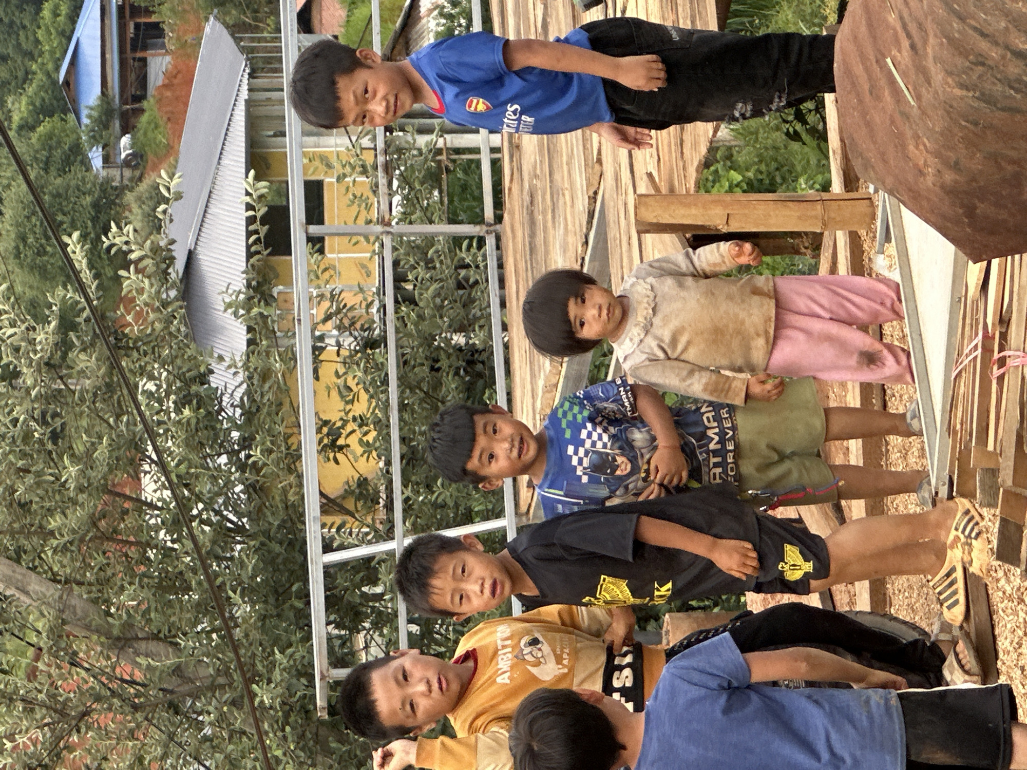 Six children, five boys and one girl, are depicted standing on and around a wooden platform in what is identified as Xín Vàng, Vietnam. In the foreground, from left to right, a boy with his back partially to the camera wears a blue t-shirt and black shorts. Next to him, a taller boy with short dark hair wears a yellow long-sleeved shirt with "AMBITION" and "APACHE" graphics, and dark pants; his left hand is raised near his head. To his right, a younger boy with short dark hair wears a black t-shirt with a yellow emblem and black shorts with a matching emblem. In front of him, a small girl with a bob haircut wears a light-colored, ruffled-collar top and pink pants, both visibly soiled. To her right, another boy wears a blue t-shirt with a Batman graphic and greenish-brown shorts. On the far right, a boy with short dark hair wears a blue Arsenal FC jersey and black pants, looking towards the camera.
The children stand on an unfinished wooden plank structure, possibly a raised platform or a construction site, with additional lumber visible in the immediate foreground. Behind the children, a grey metal railing spans horizontally across the midground. Beyond the railing, a two-story building with a yellow facade and a dark corrugated metal roof is visible through dense green foliage. Further in the background, amidst hills covered in green trees, another building with a bright blue roof is partially seen. Overhead, thin black lines, likely utility wires, cross the upper portion of the frame. The lighting is diffused, suggesting an overcast day.