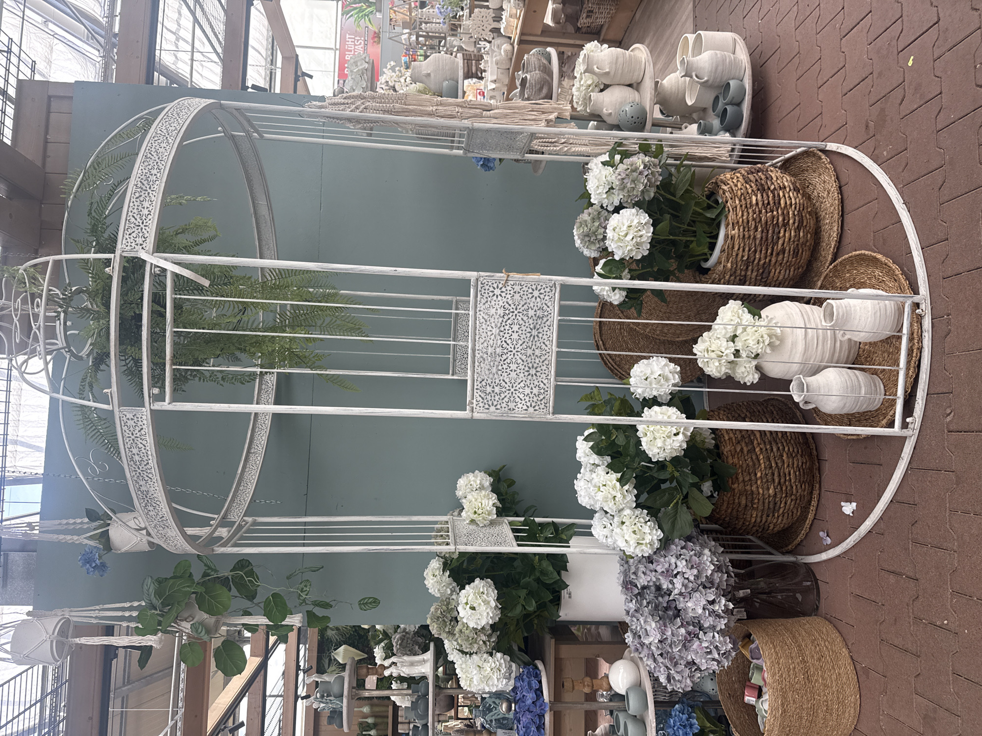 The image depicts a retail display of artificial flowers and decorative pottery in a garden center or flower shop. The primary subject is a large, white, ornate birdcage-like structure serving as a backdrop. Within and around this structure are arrangements of white and purple hydrangeas, some in woven baskets and white ceramic pots. Several white ceramic vases and jugs of varying sizes are placed on woven mats at the base of the display. Hanging plants in macrame holders are suspended from above. In the background, other retail items, including shelves with pottery and decorative objects, are visible. The flooring is made of red brick pavers. The scene is lit by natural light, suggesting an indoor or covered outdoor space. The location is identified as a garden center in Fritzlar, Germany, based on a partial sign visible in the background which includes the word "FRITZLAR". There are no people or animals visible in the image.