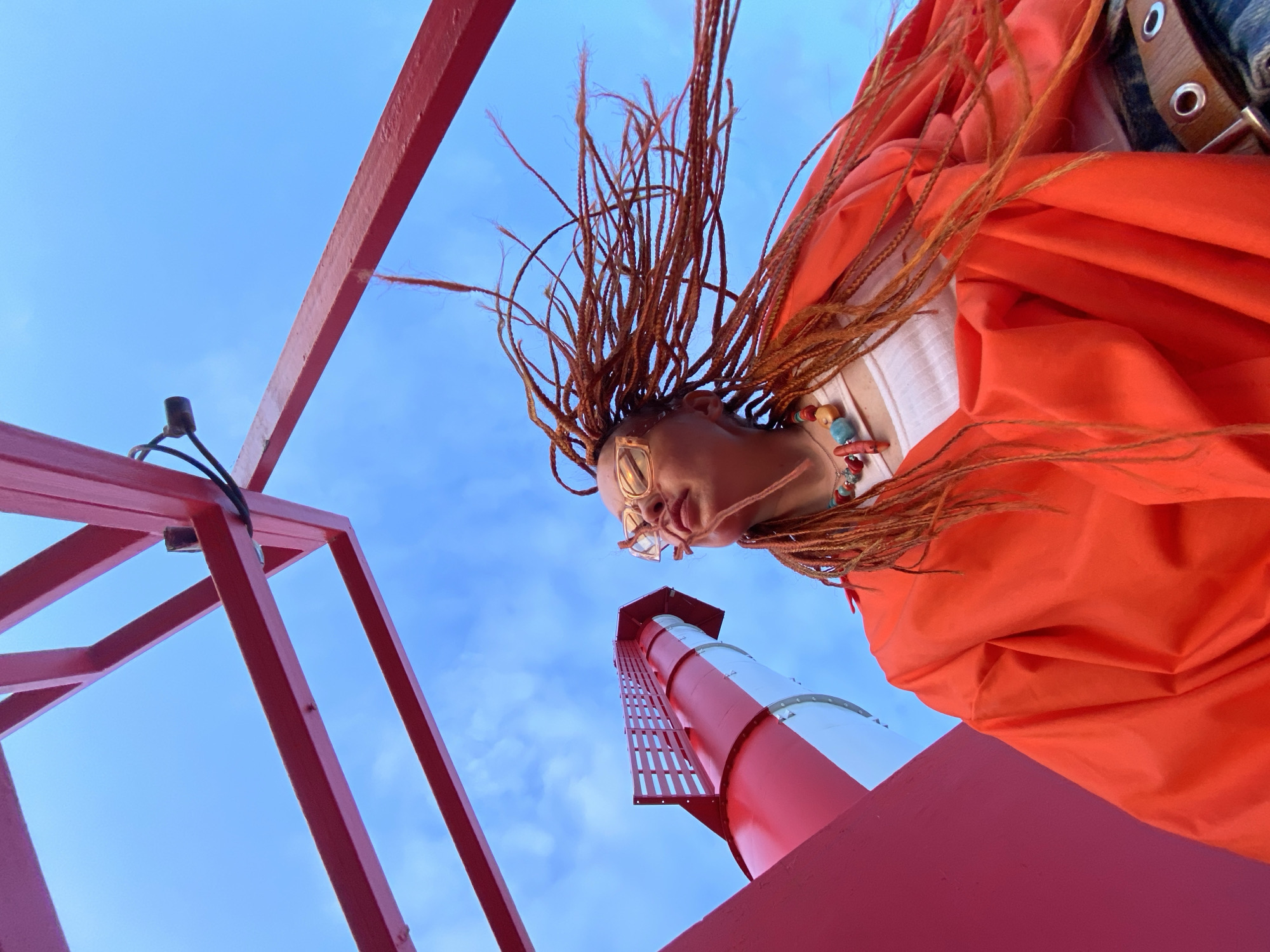 A low-angle shot captures a person from the chest up, looking upwards against a bright blue sky. The person has long, braided auburn hair, is wearing a vibrant orange jacket over a white top, and a beaded necklace. They are also wearing glasses. In the background, a red metal structure is visible, as is a large, red and white cylindrical structure that appears to be part of a tower or pipe. The image was taken in Sochi, Russia.