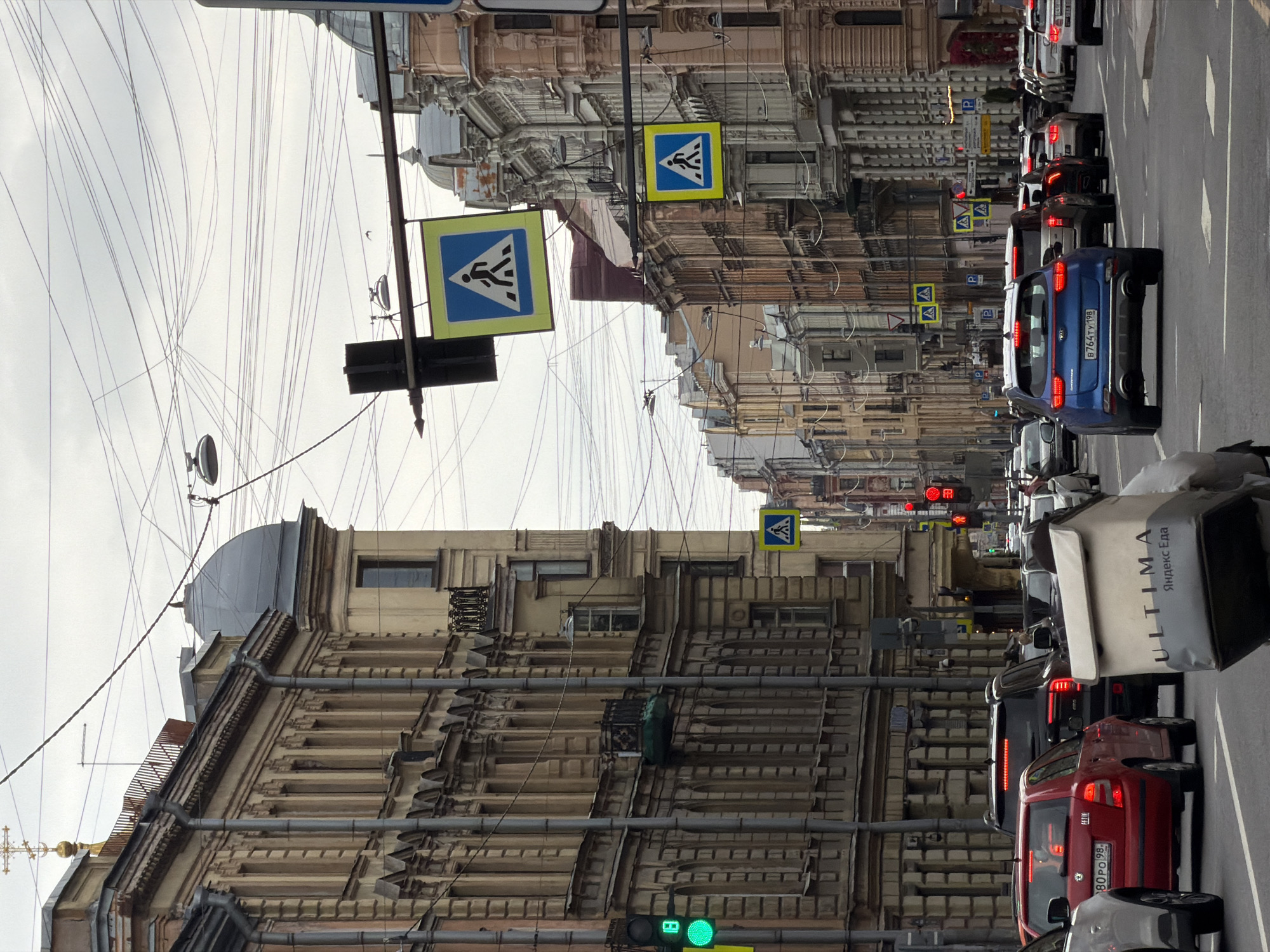 The image captures a street scene in Saint Petersburg, Russia, characterized by heavy traffic and densely packed buildings. The shot is taken from street level, looking down a narrow avenue lined with ornate, historical buildings. Overhead, a complex network of power lines crisscrosses the sky. Pedestrian crossing signs are prominently displayed, indicating designated areas for crossing the street. Cars are densely packed along the road, with visible tail lights suggesting stopped or slow-moving traffic. The light conditions appear overcast, casting a subdued light on the cityscape. The presence of delivery services can be seen in the vehicles, adding to the urban hustle.