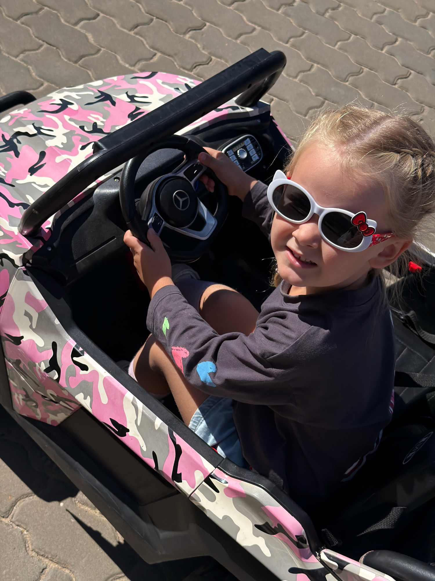 A young Caucasian girl with blonde hair in braids is sitting in a pink and gray camouflage toy electric car. She is wearing white cat-ear sunglasses with red bows on the sides, a dark gray long-sleeved shirt with colorful designs on the sleeve, and light blue shorts. She is holding the steering wheel of the car, which has a Mercedes-Benz logo. The car is parked on a paved surface made of interlocking concrete blocks, commonly found in urban areas of Kamyshin, Russia. The lighting suggests it is daytime, with clear sunlight casting shadows on the ground.