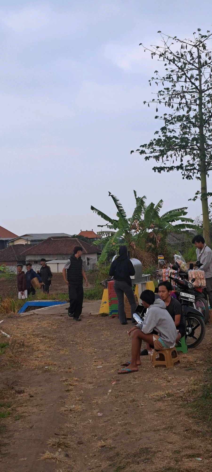 In Sumber Gedang, Indonesia, on a cloudy day, a group of young men and women are gathered outdoors in a rural setting. The ground is dirt with sparse, dry grass. Behind the people are buildings with terracotta tiled roofs, characteristic of Indonesian architecture. A large tree with hanging pods dominates the right side of the frame, with banana trees visible closer to the ground.
In the foreground, three young men are seated on small stools, with one man holding a cigarette and another looking towards the right. A motorcycle is parked behind them, loaded with bags of snacks.
Further back, a young woman wearing a black hijab and pants stands near a metal apparatus. To her left, a young man in a black vest and pants walks away from the camera. Behind him, three more young men are standing; one carries a woven basket.
The overall scene appears to be a casual gathering or break, possibly during an activity or event. The environment suggests a blend of rural life and everyday activities.