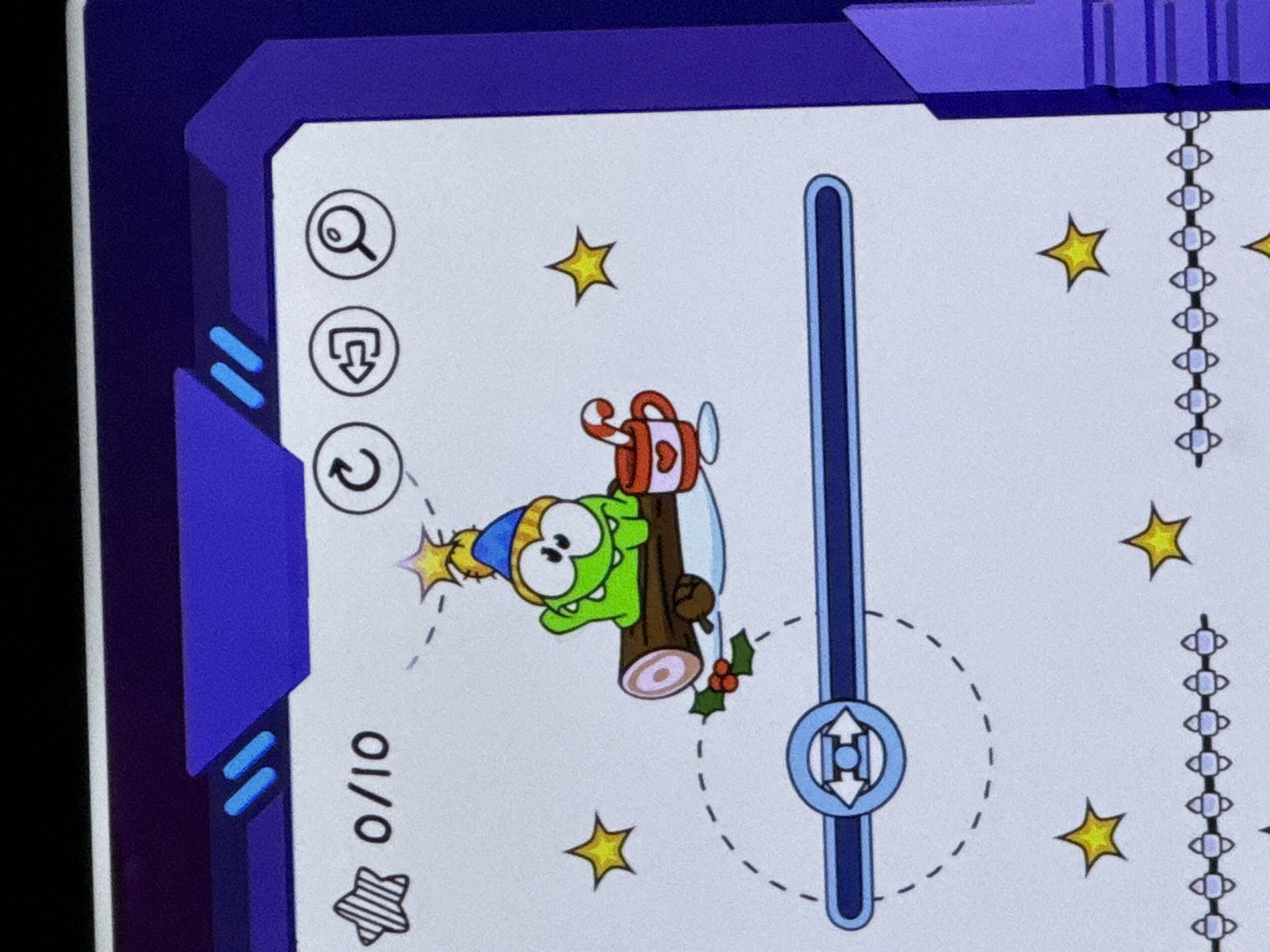 This is a screenshot of a mobile game, likely "Cut the Rope" given the character. The game displays a cartoon frog-like character wearing a blue and yellow winter hat, sitting on a log. Next to the frog is a mug with a candy cane in it. The background is white with dashed lines and yellow stars scattered around. At the top left, there's a score indicator showing "0/10" next to a striped star. To the right of the score are three circular icons: a circular arrow, a backward arrow, and a magnifying glass. A blue progress bar is visible near the bottom of the screen. The image appears to be taken on a mobile device, indicated by the purple border around the screenshot. There is no information in the image that places it in the city of Cahul, Moldova.