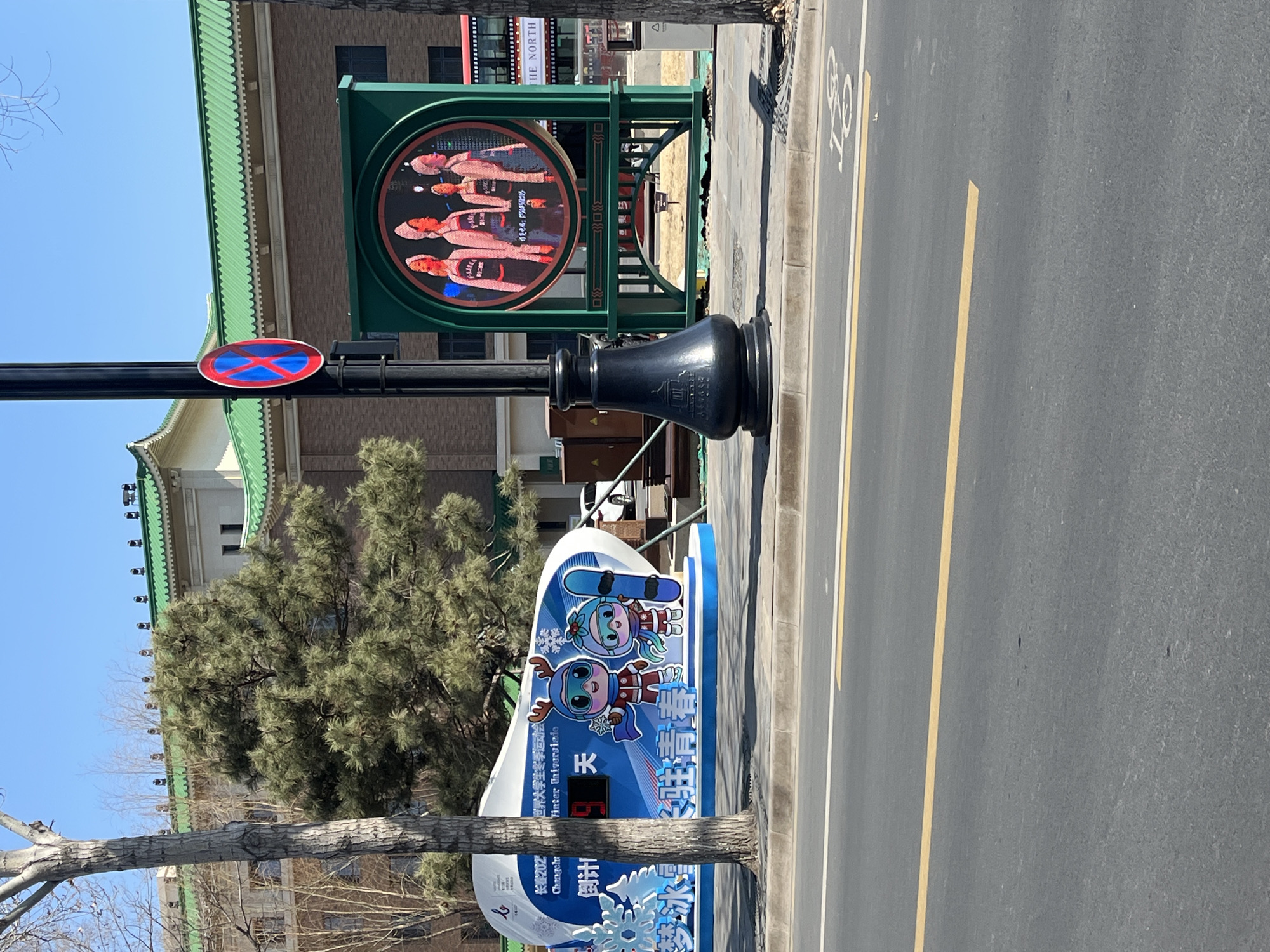 Here is a detailed description of the image:
The image shows a street scene in Changchun City, China. The camera is positioned on the side of the road. The street is divided by a yellow line and has a white bicycle lane marking on the right side.
The scene includes several prominent signs. A green decorative sign with a circular display showing athletes in red outfits is mounted on a black lamp post. The lamp post also has a “no parking” sign attached to it. A large blue and white sign promoting the 202 Winter Universiade in Changchun is positioned next to the street. This sign features cartoon mascots and includes a countdown timer with the number 5 displayed. Text in both Chinese and English (“Winter Universiade”) is visible on the sign.
A building with a traditional green-tiled roof is in the background, partially obscured by a tree. The overall environment suggests a clear, sunny day. Another sign that reads "THE NORTH" is visible in the background.